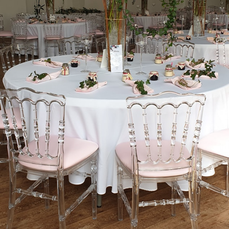 Table ronde décorée avec une nappe blanche, verres à vin, assiettes avec serviettes rose, petites caissettes avec crème et confiture, feuilles vertes et branches, dans une salle de réception élégante.