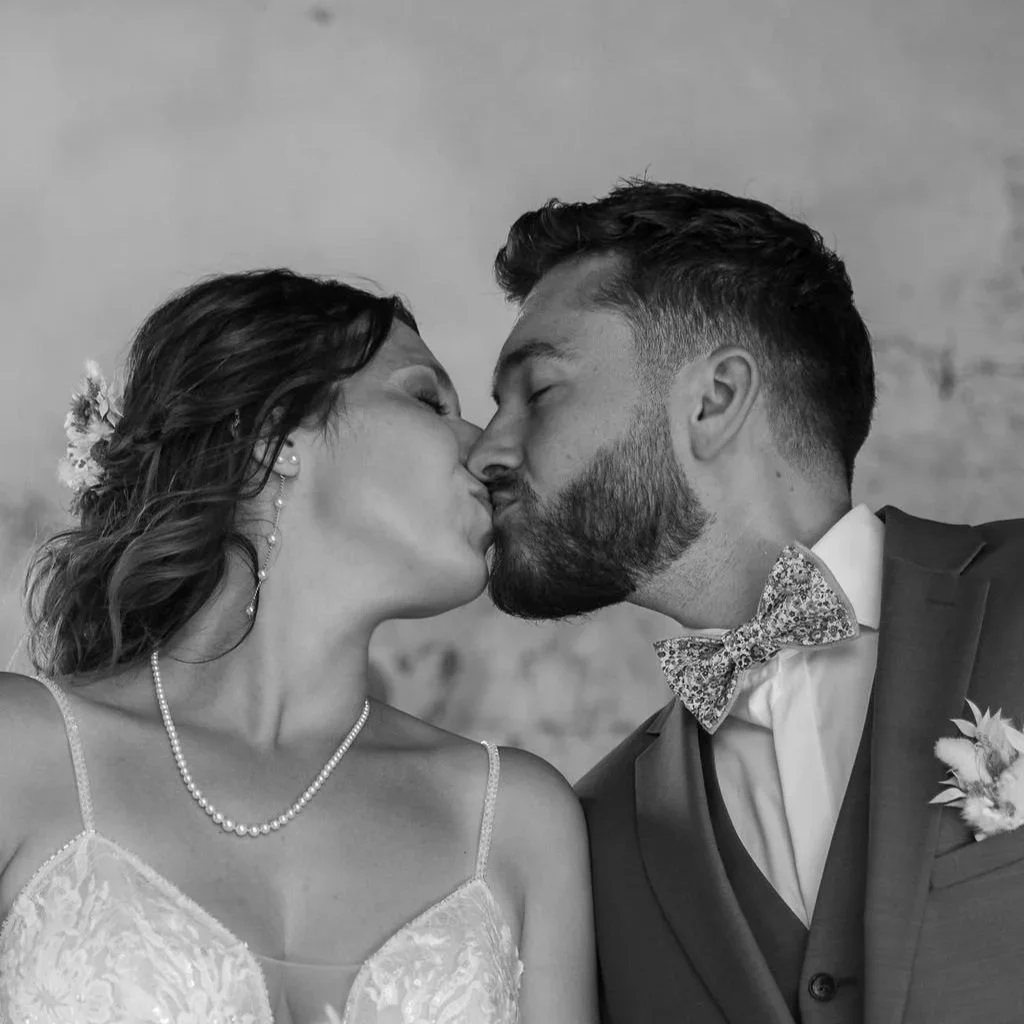 Un couple habillé en mariage s'embrassant, la femme porte une robe à bretelles et un collier de perles, le homme porte un costume avec nœud papillon.