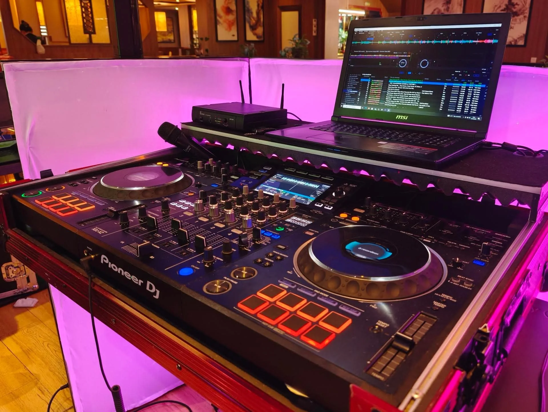 Une table de DJ avec un contrôleur Pioneer DJ, un ordinateur portable affichant un logiciel de mixage, et un micro, dans une ambiance de lumière rose.