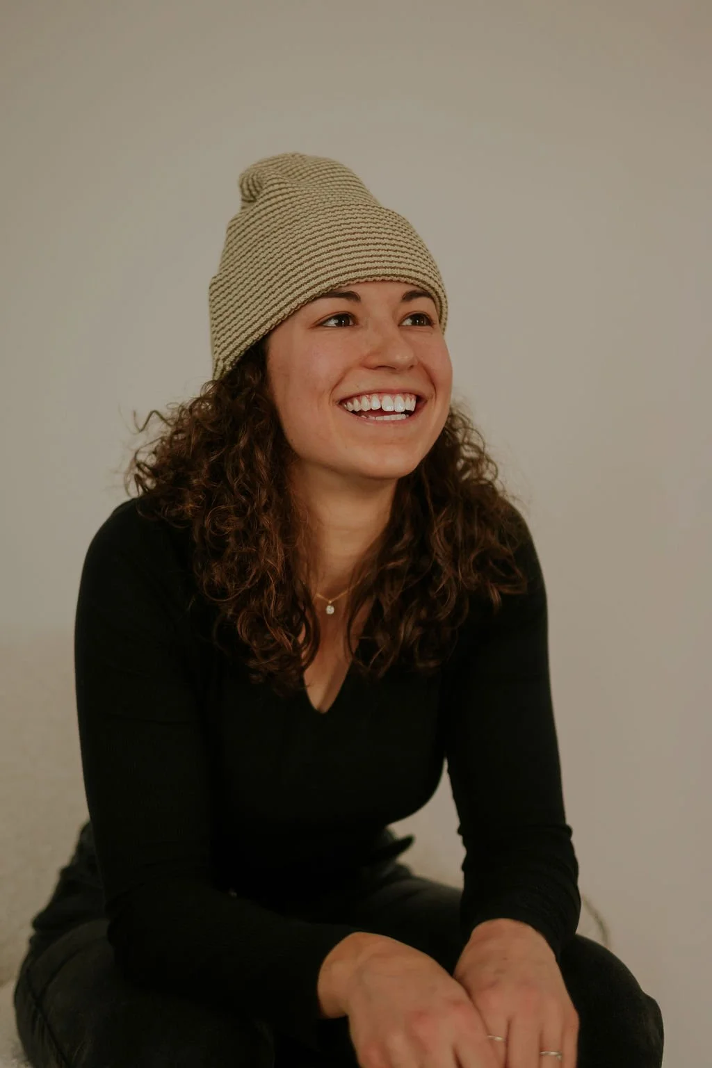 Une femme souriante avec des cheveux bouclés, portant un bonnet beige à rayures et un haut noir, assise contre un mur blanc.