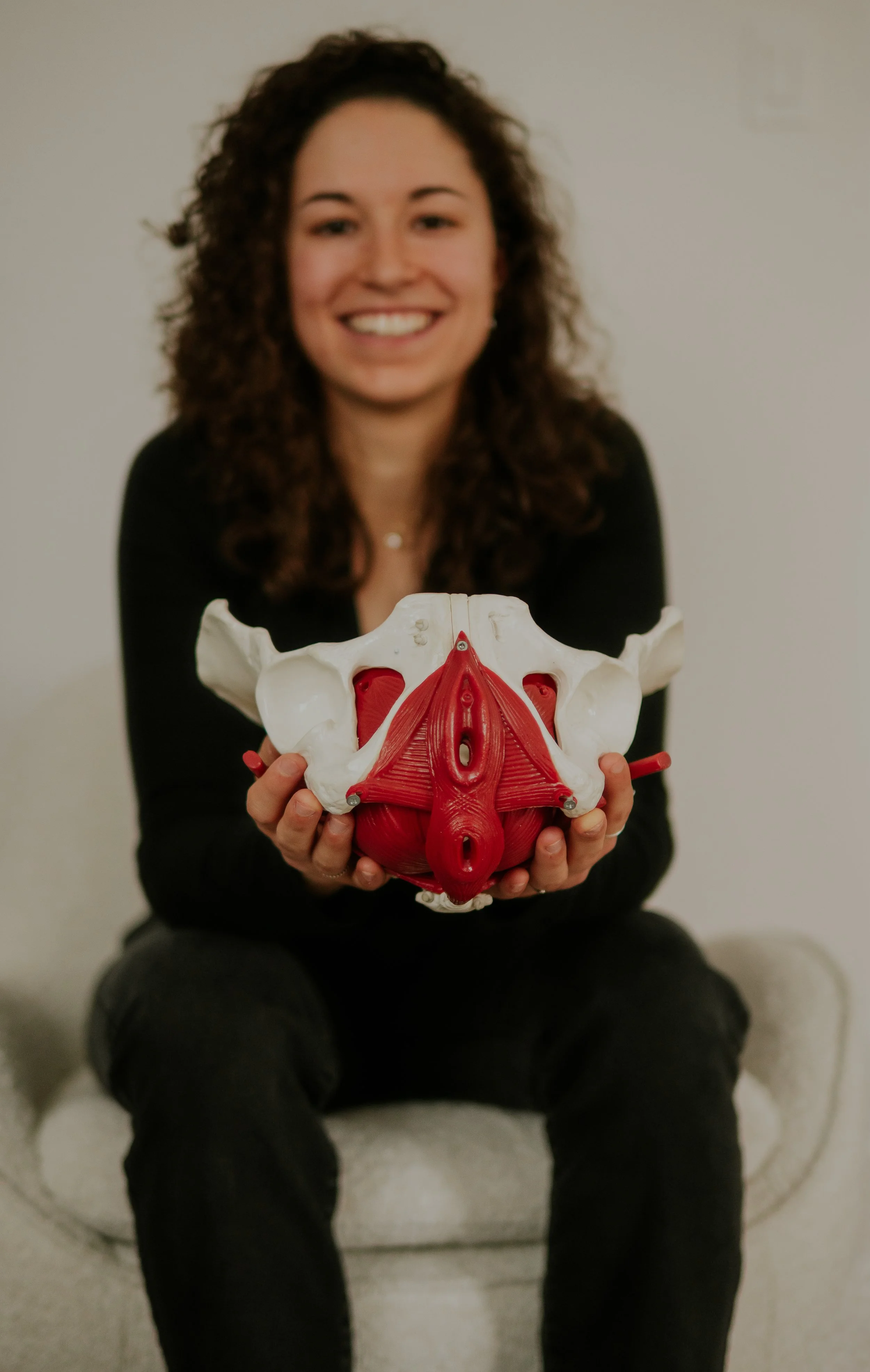 Woman with curly hair smiling and holding a detailed anatomical model of the pelvic region.