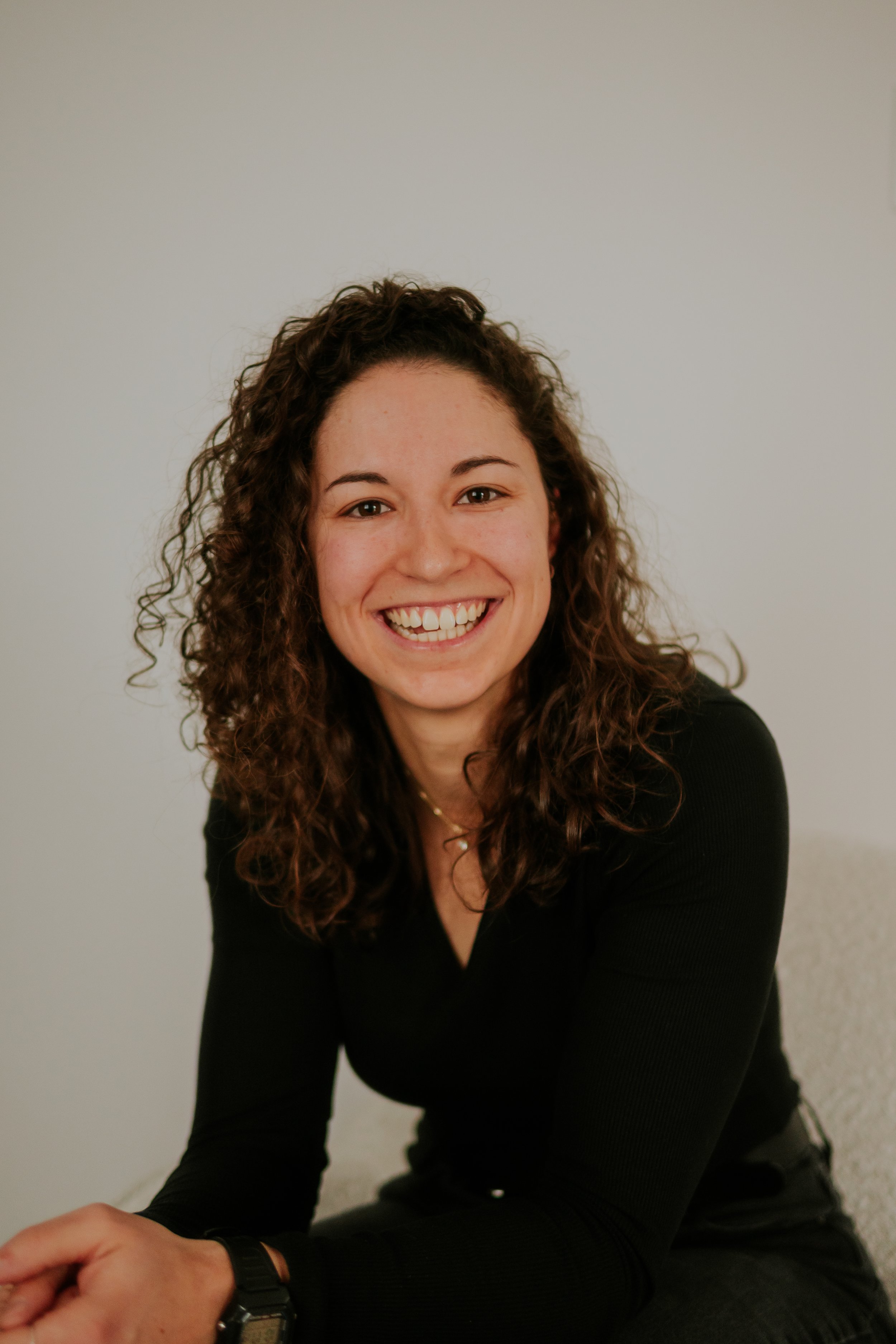 A woman with curly brown hair smiling and sitting in front of a plain white wall, wearing a black long-sleeve top.
