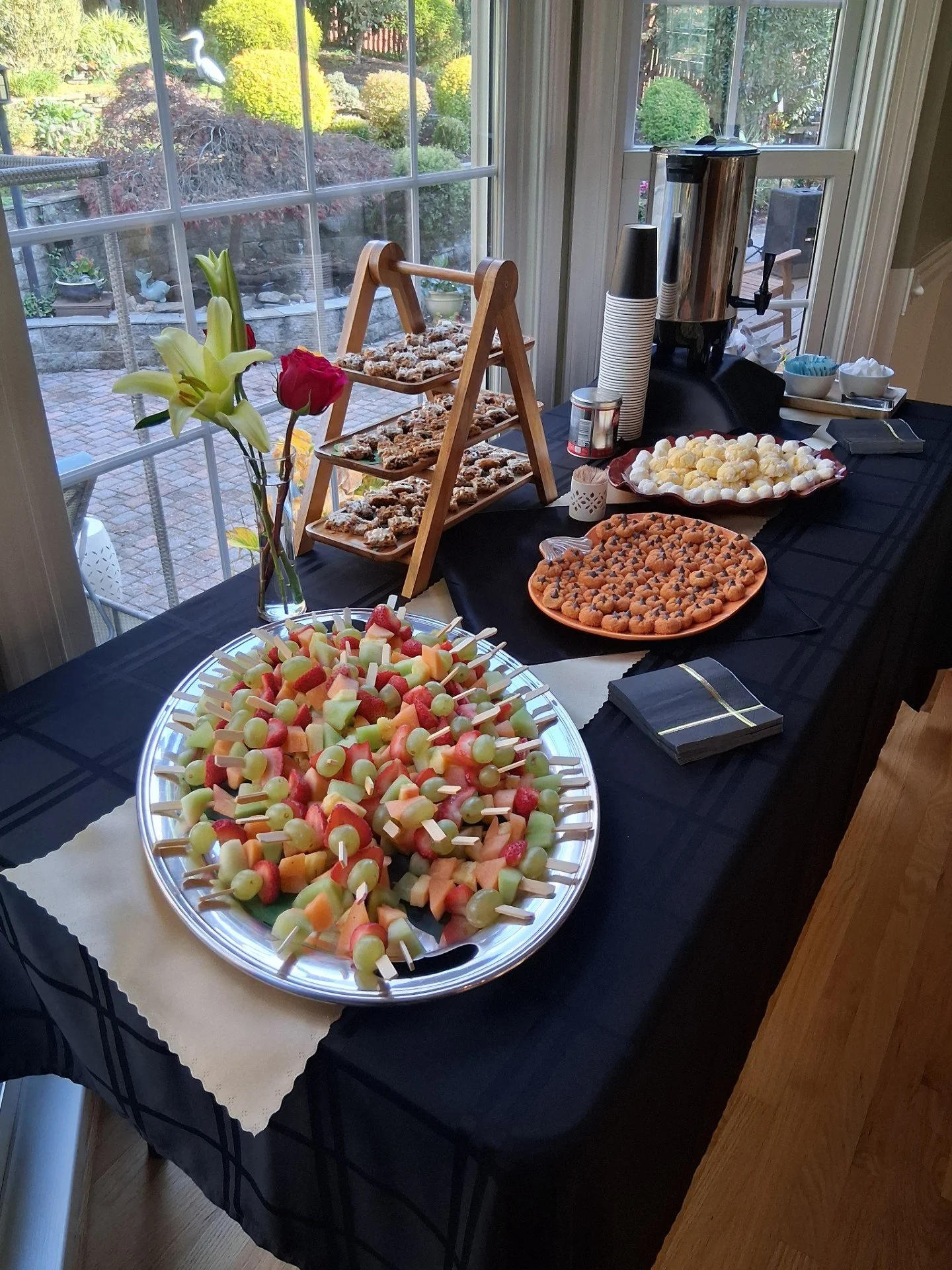 A table with a black tablecloth set for a gathering, featuring a platter of assorted fruit skewers, a vase with lilies and roses, multiple plates with cookies, a hot beverage dispenser, cups, and napkins near a window with a garden view.