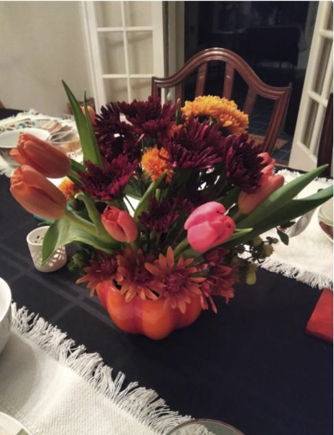A vibrant floral centerpiece with tulips, daisies, and chrysanthemums in a pumpkin-shaped container on a dining table.