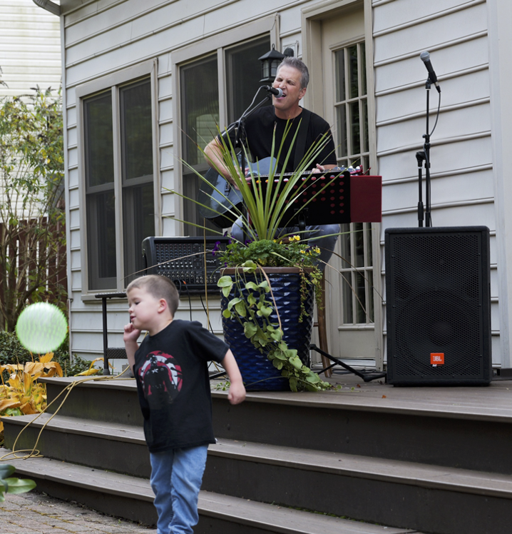Man performing a musical act on an outdoor stage with a microphone and guitar, decorated with a large potted plant, while a young boy in front plays with a ball outdoors.