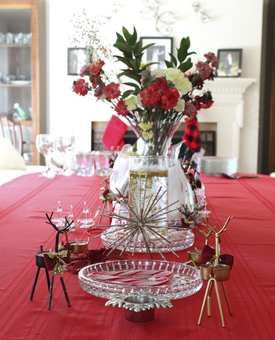 A festive table decorated with a red tablecloth, a tall glass vase with pink and white flowers, and gold reindeer and star ornaments, likely for a holiday celebration.