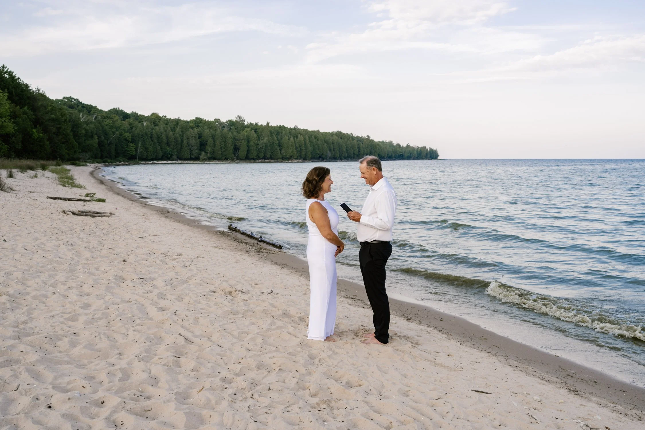 A couple getting married on a beach, with the groom reading vows from a mobile phone, while the bride watches and smiles.