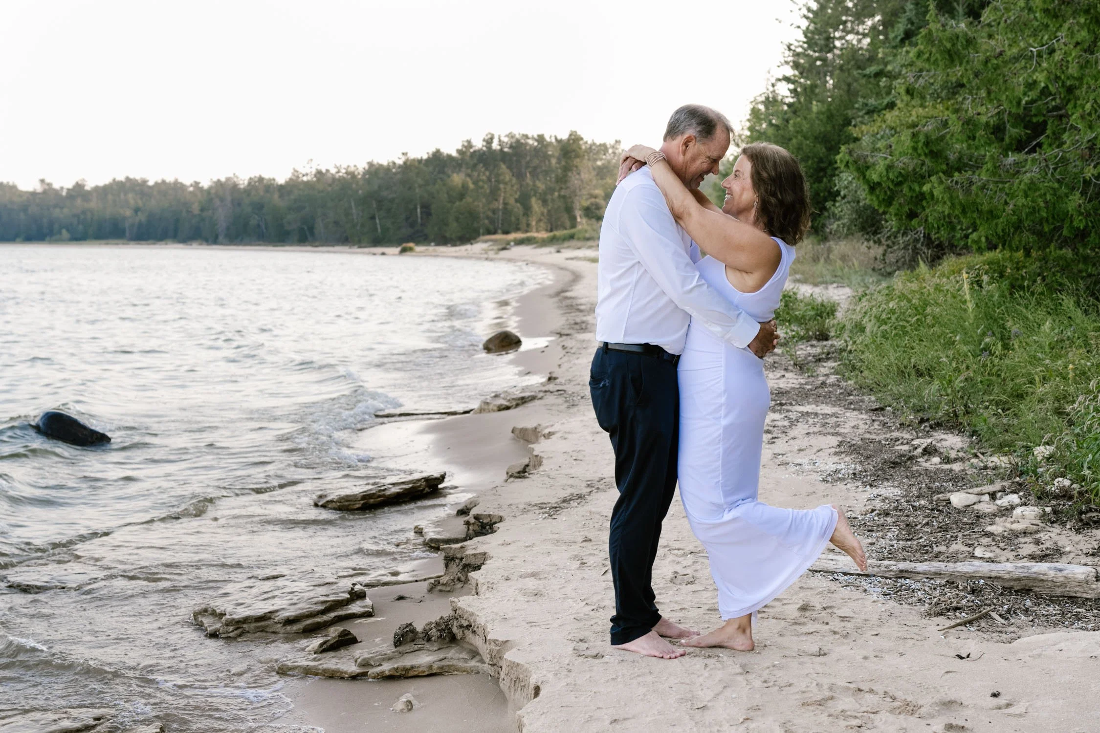 A happy couple embraces on a sandy beach by the water, holding each other and smiling.