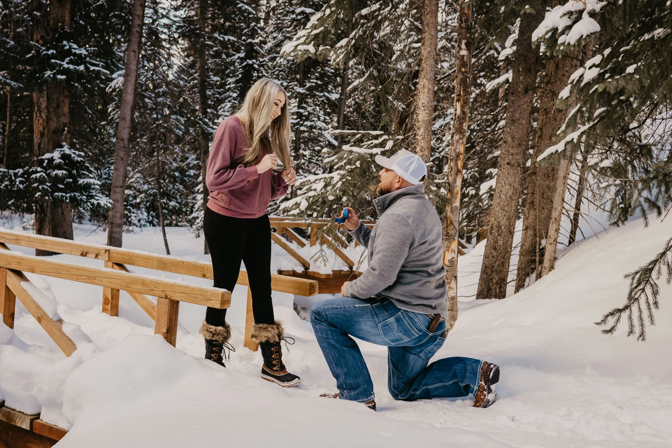 A man is proposing marriage to a woman on a snowy outdoor wooden staircase in a forest, with snow-covered trees in the background.