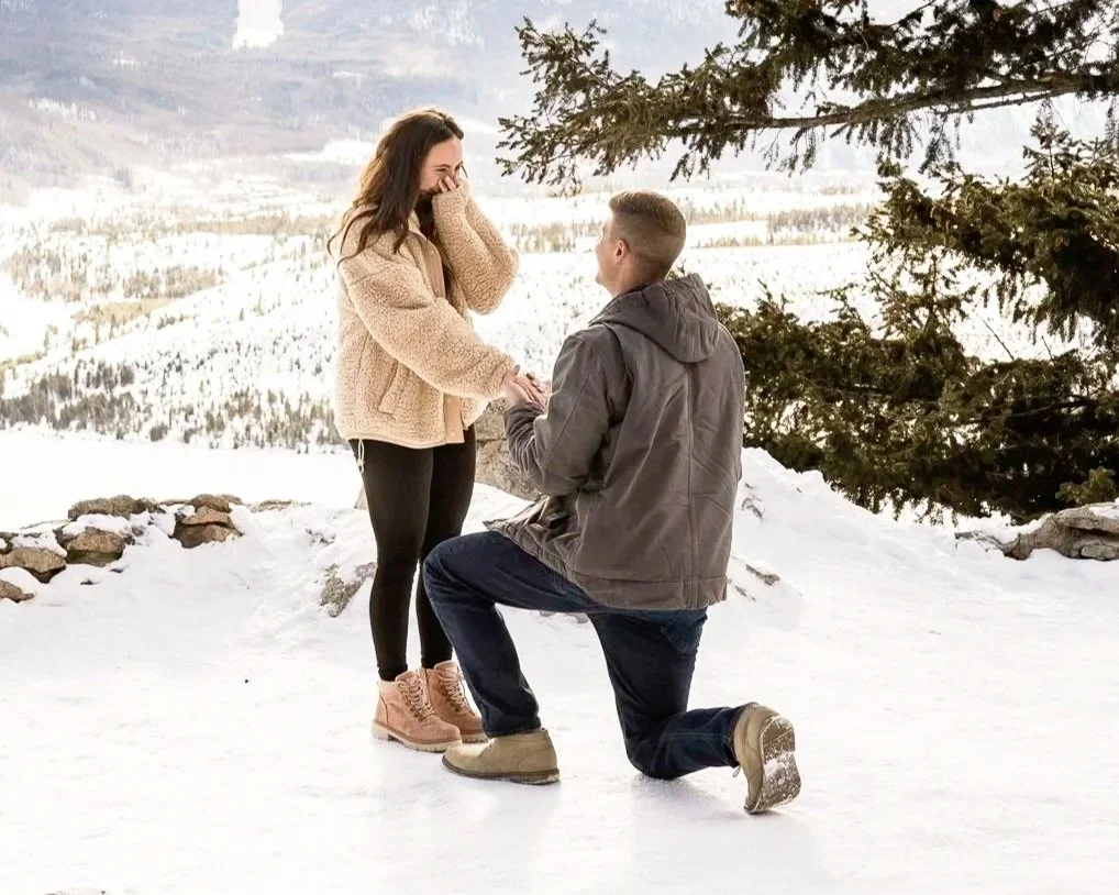 A man proposing to a woman in a snowy outdoor setting with mountains and trees in the background.