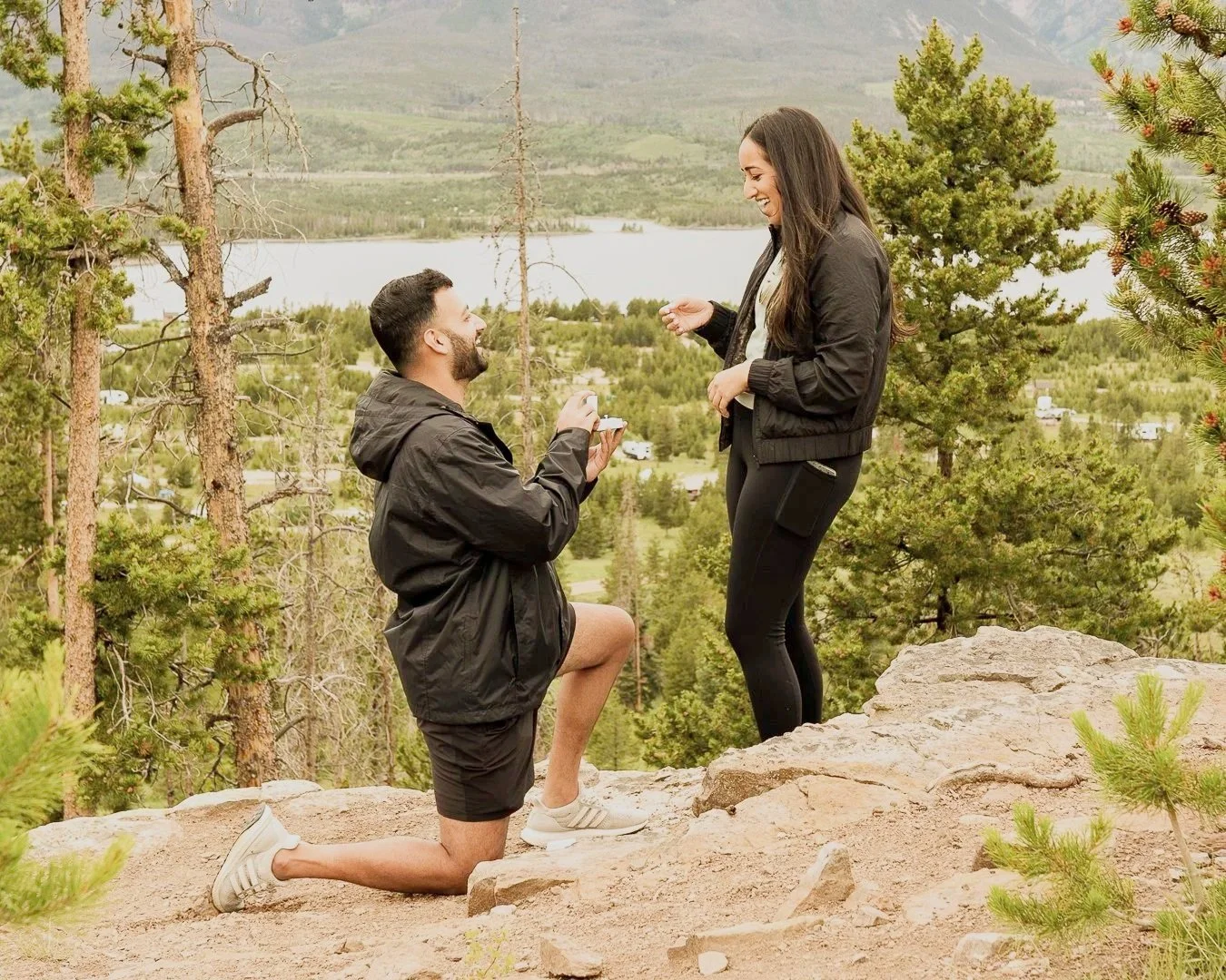 A man proposing to a woman on a rocky overlook surrounded by trees with a lake and mountains in the background.