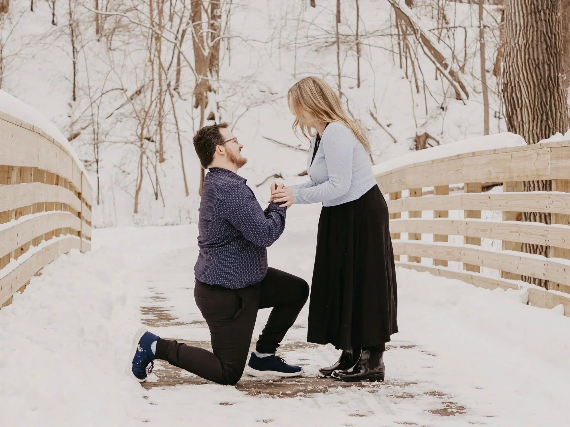 A man is kneeling on one knee on a snow-covered wooden bridge, proposing to a woman in a wintry forest. The woman is standing and smiling while holding his hands.