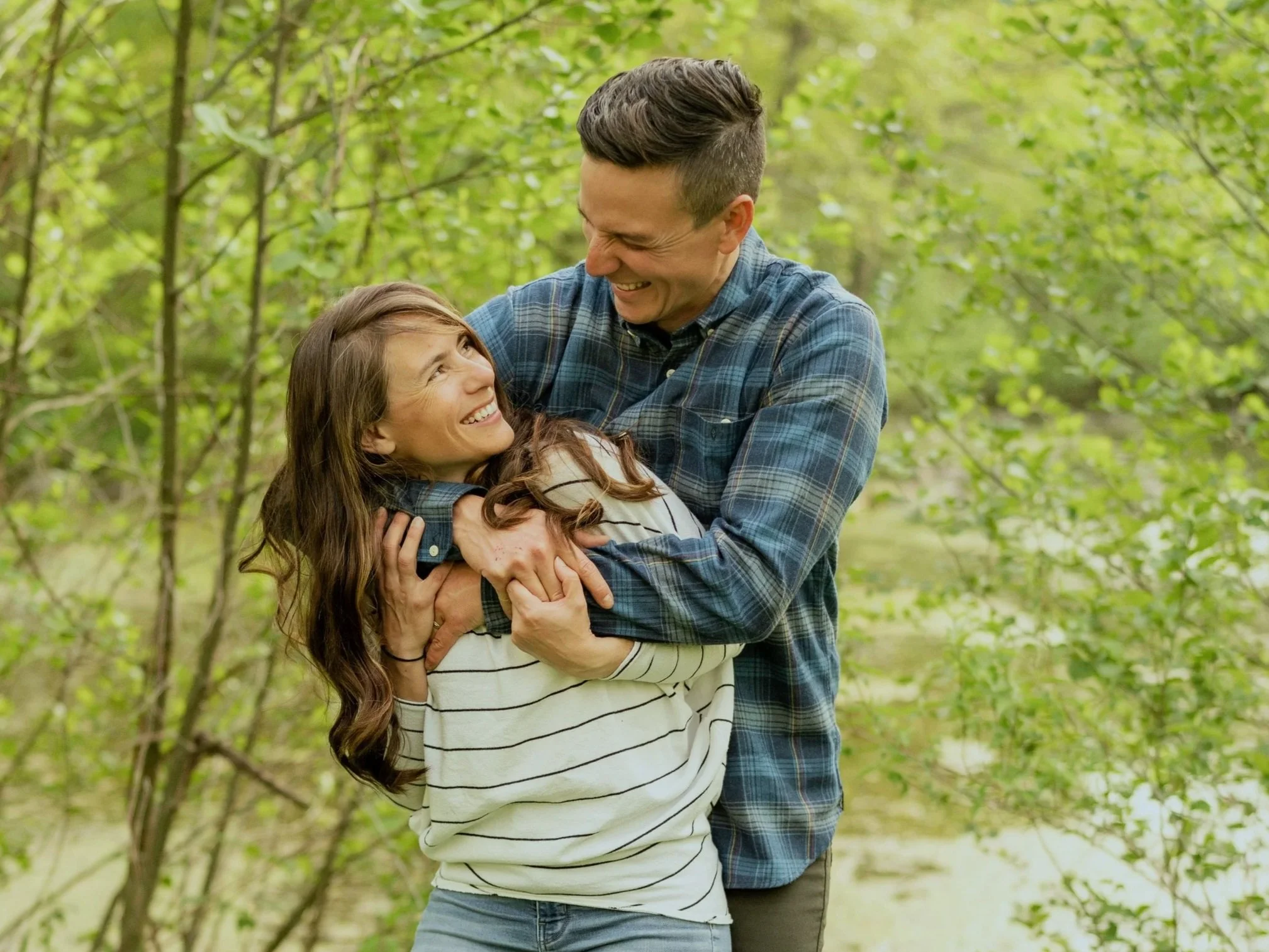 A couple joyfully embracing in a forested area with green leaves.