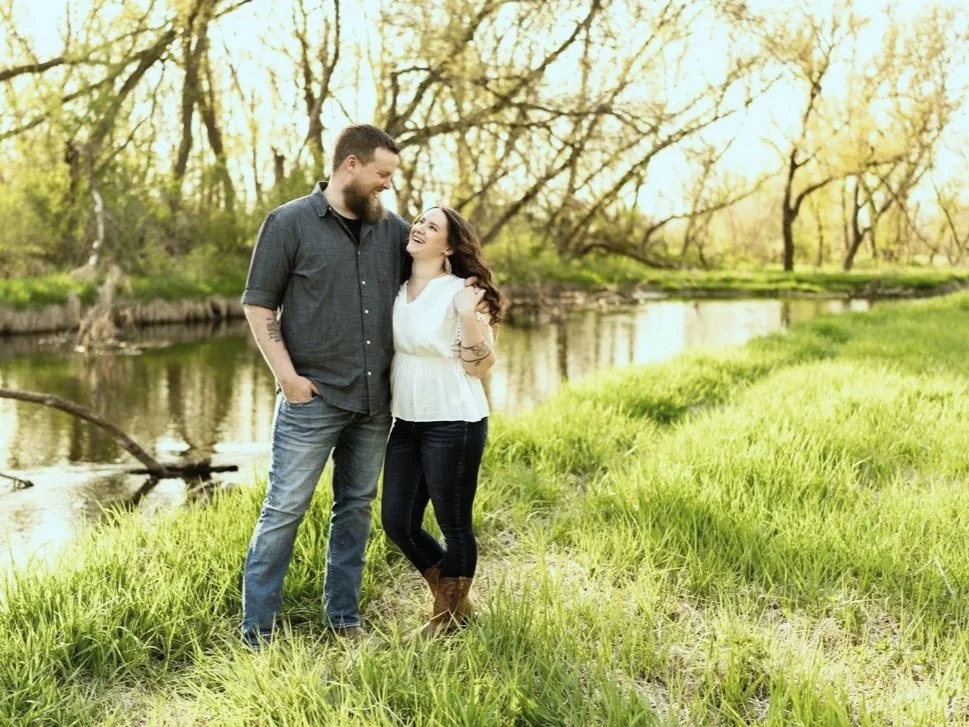 A couple standing in a grassy area near a river, smiling and looking at each other, surrounded by trees with yellowish leaves.