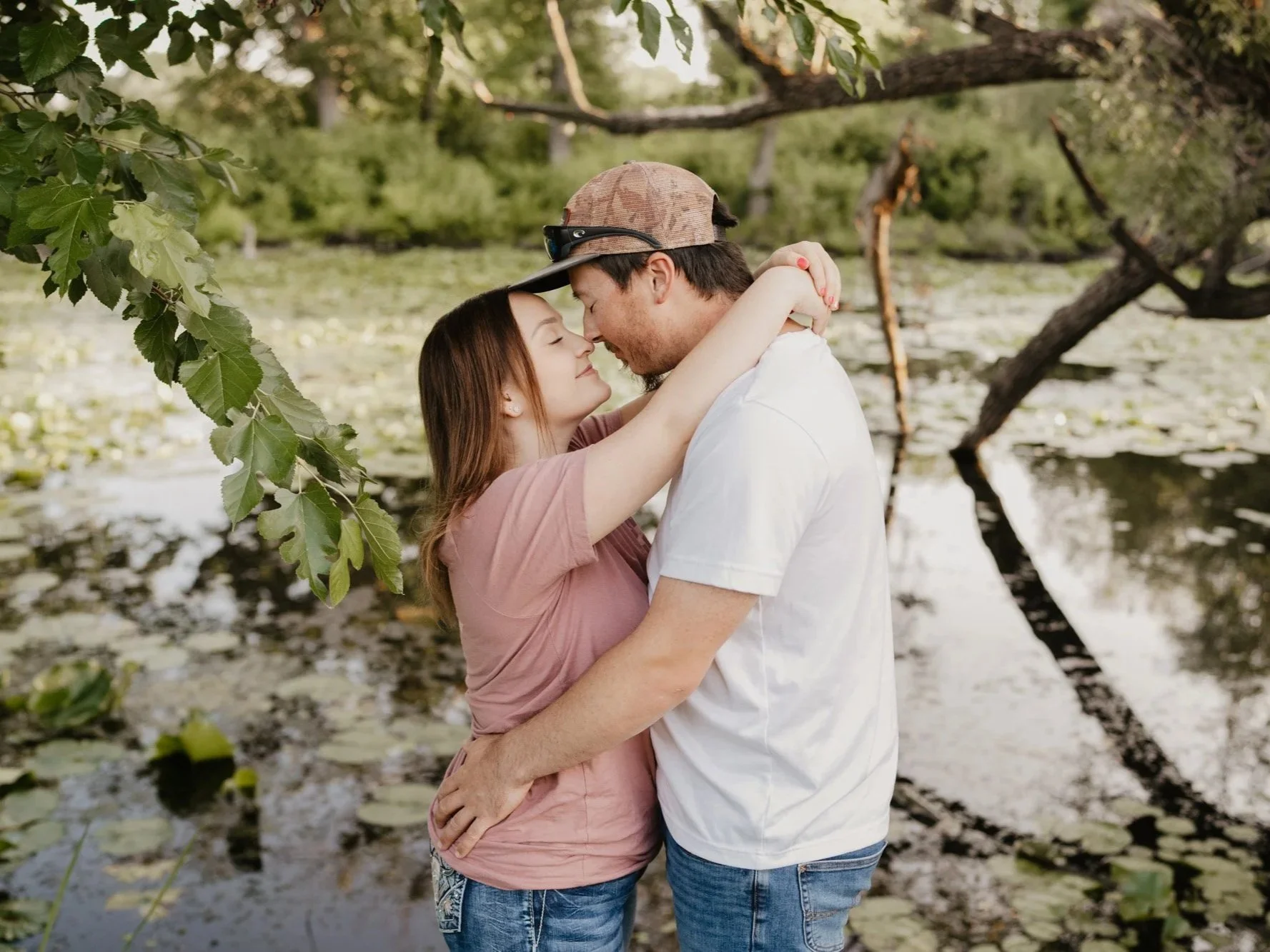A couple embraces by a pond with lily pads and a bent tree branch overhanging the water, surrounded by green foliage.
