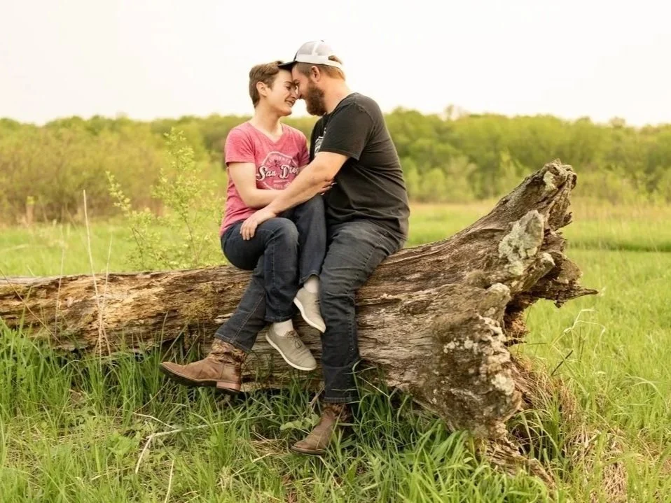 A couple sitting on a fallen tree in a grassy field, touching foreheads and smiling at each other.