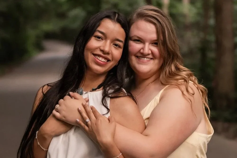 Two women hugging outdoors, smiling, with a blurred background of trees and a path.