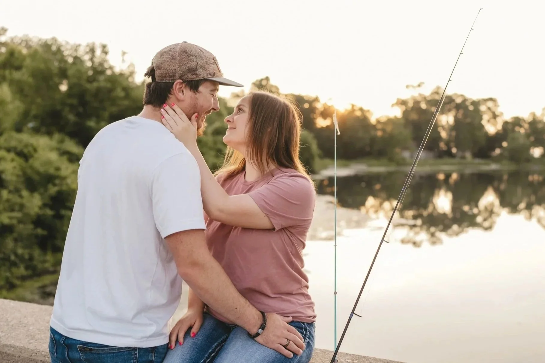 A couple stands close by a lake during sunset, with fishing rods nearby, gazing into each other's eyes and smiling.