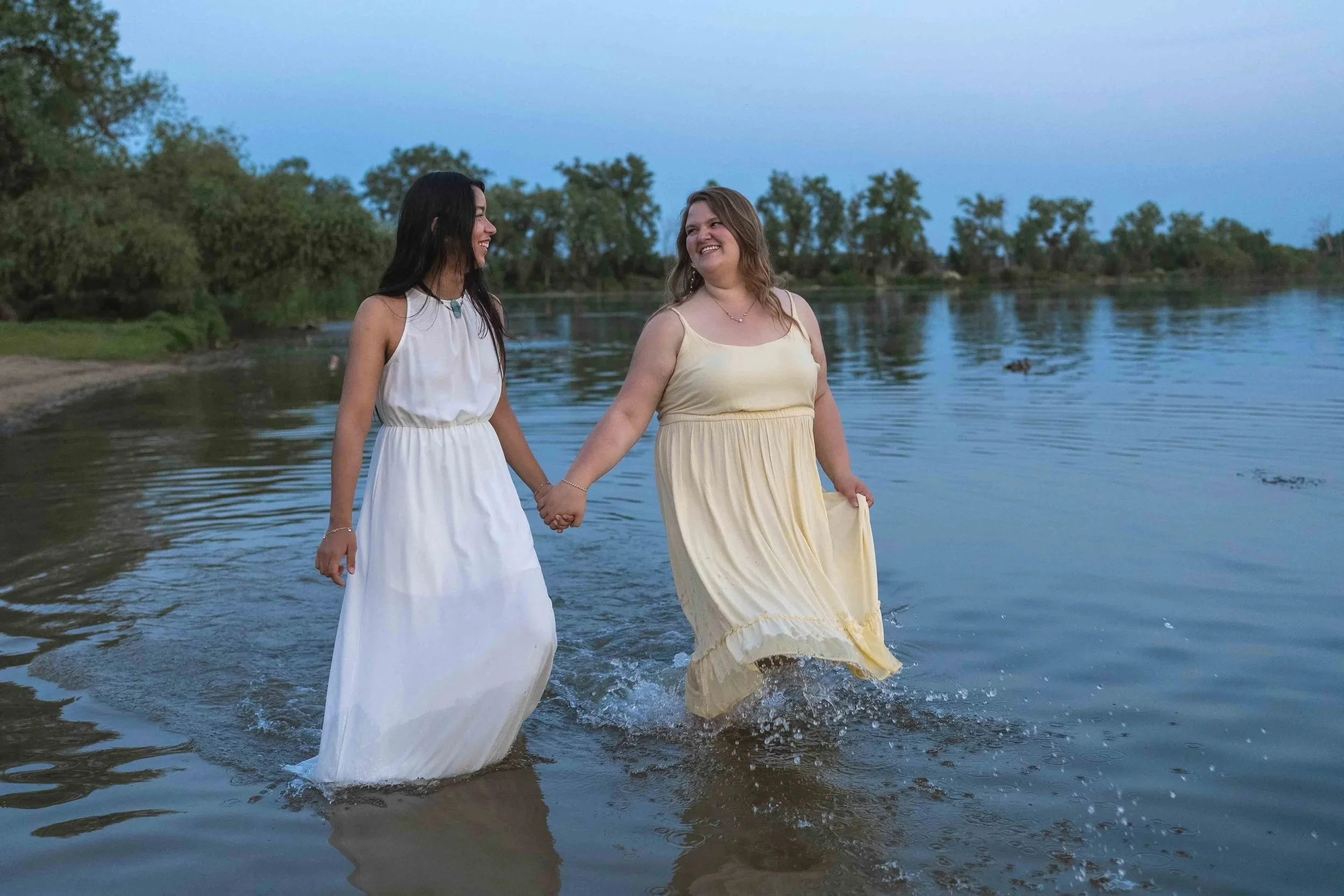 Two women in yellow and white dresses holding hands and walking in shallow water at a lake during dusk.