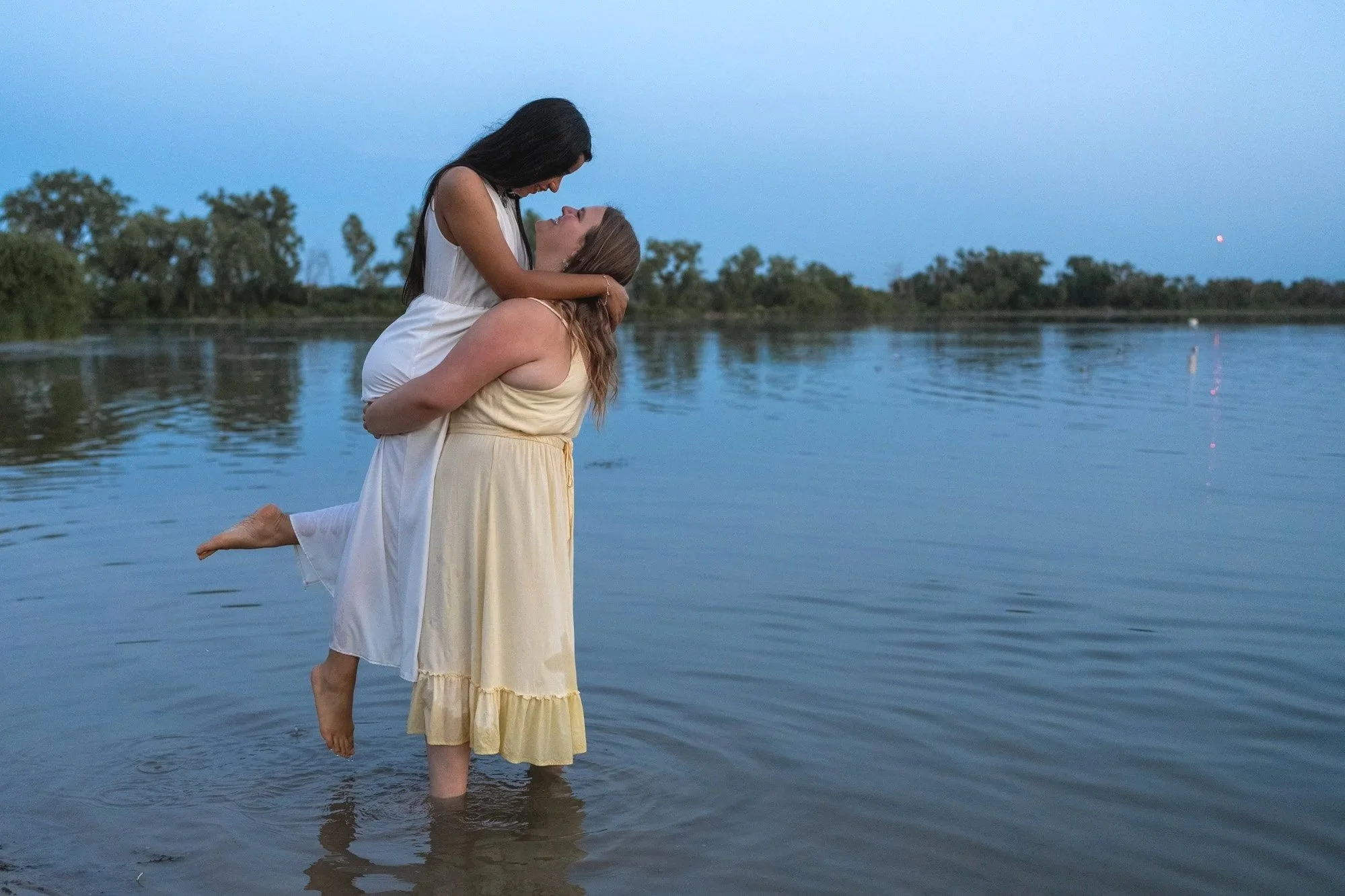 Two women in dresses embracing in a lake at dusk, with trees on the horizon and calm water.