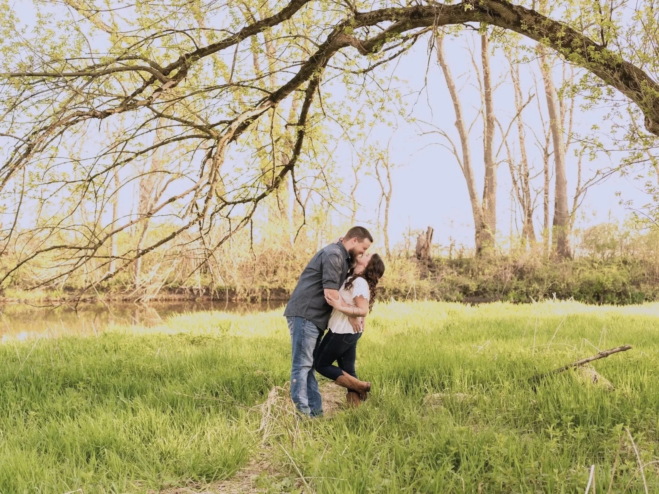 A couple kissing in a grassy area near a small water body with trees in the background.