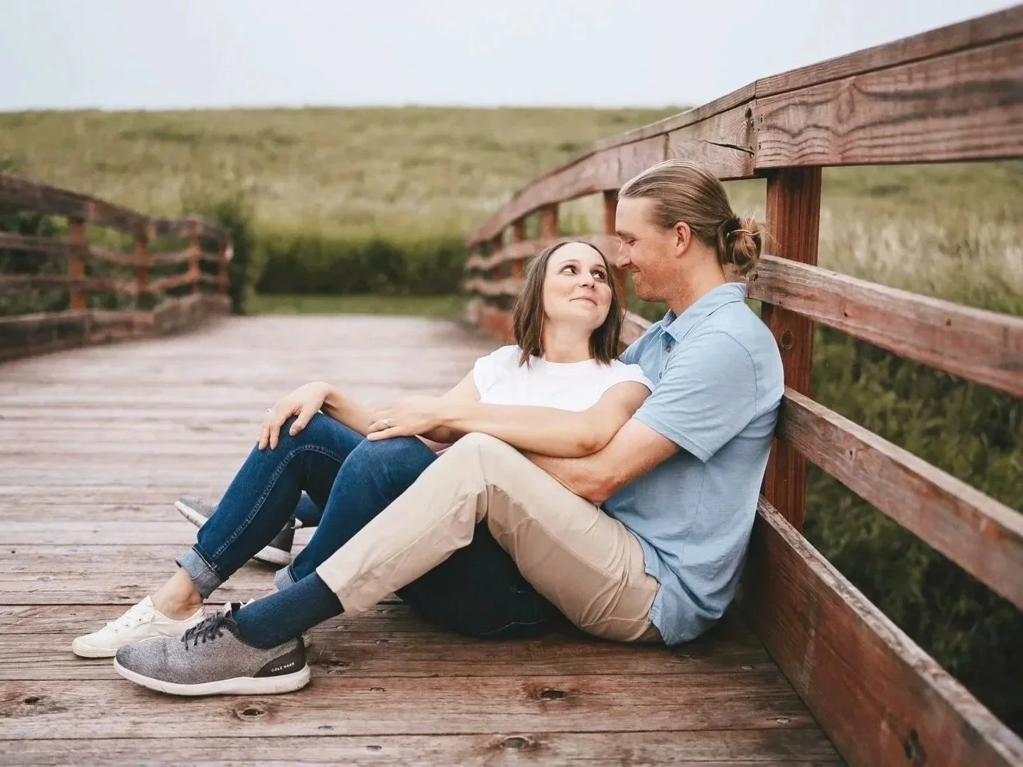 A couple sitting on a wooden bridge, facing each other lovingly, in an outdoor setting with green fields in the background for a beautiful engagement image.