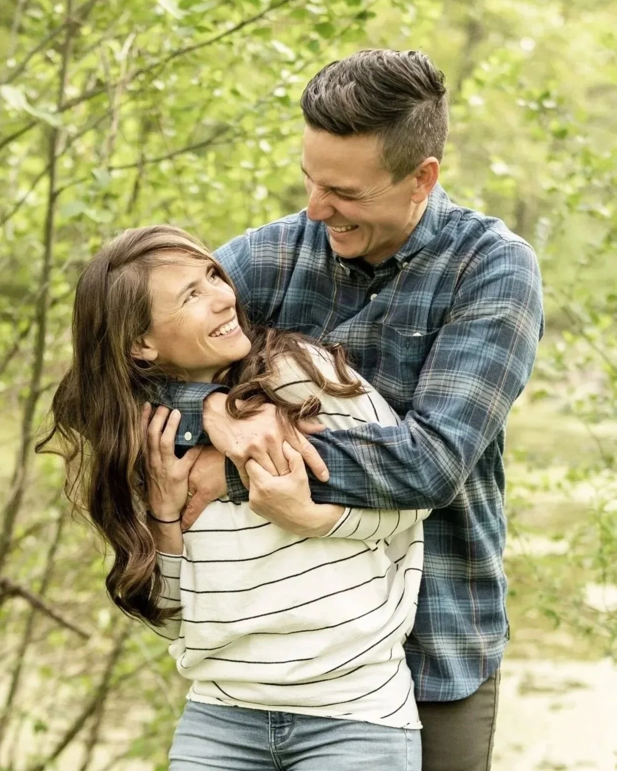 A man and woman are smiling and playing together outdoors in a wooded area. The man is hugging the woman from above, and they are looking at each other happily.