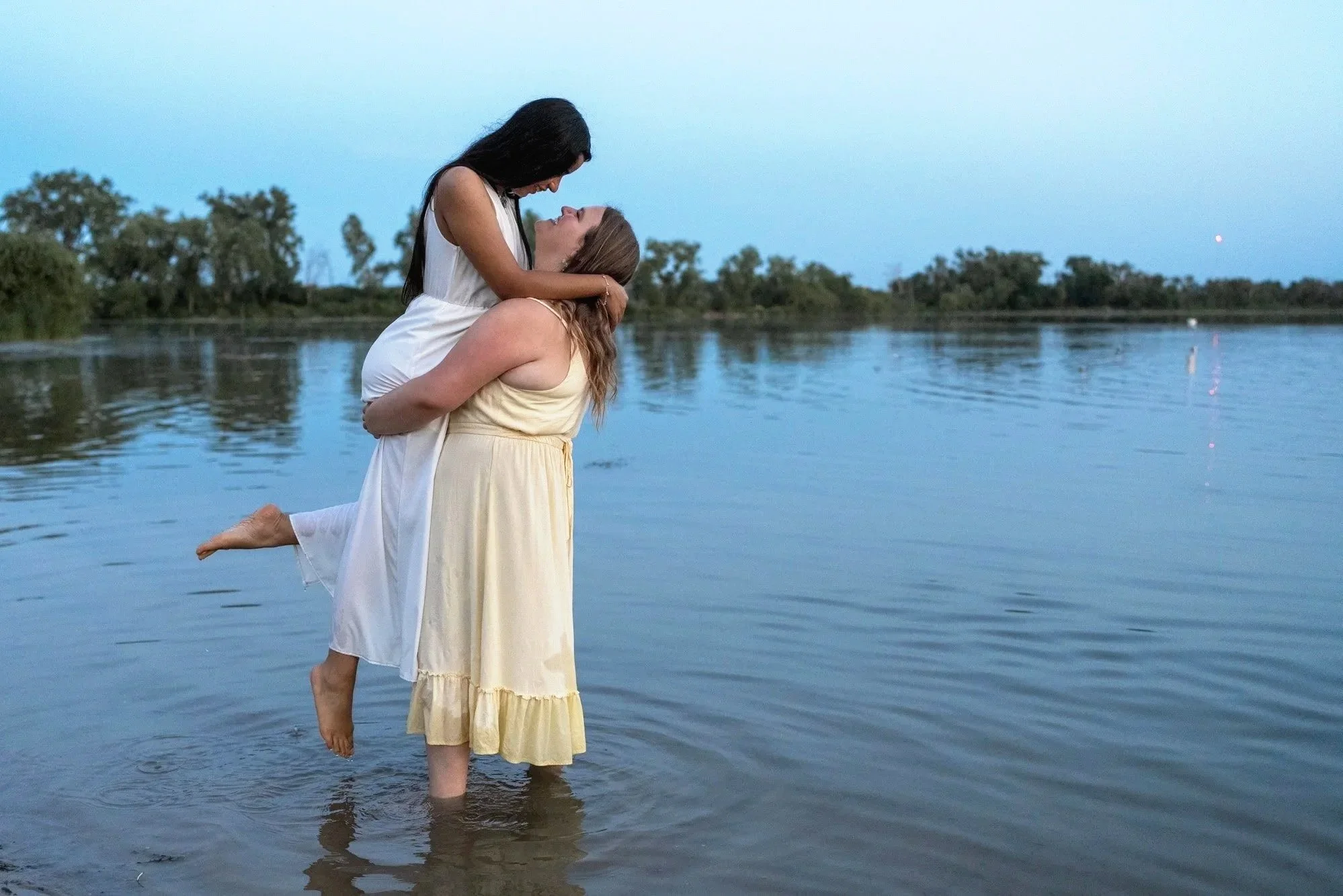 Two women in dresses embracing in a lake at dusk, with trees on the horizon and calm water.