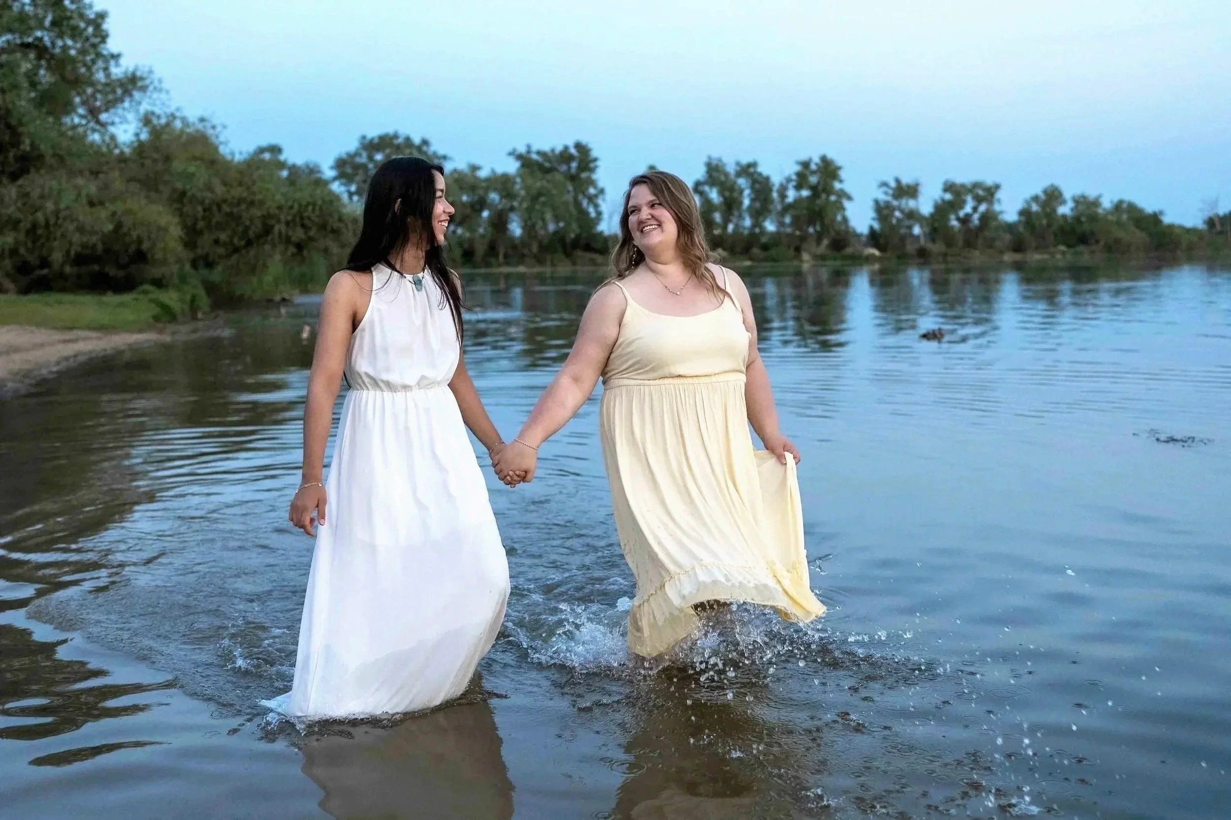 Two women in yellow and white dresses holding hands and walking in shallow water at a lake during dusk.