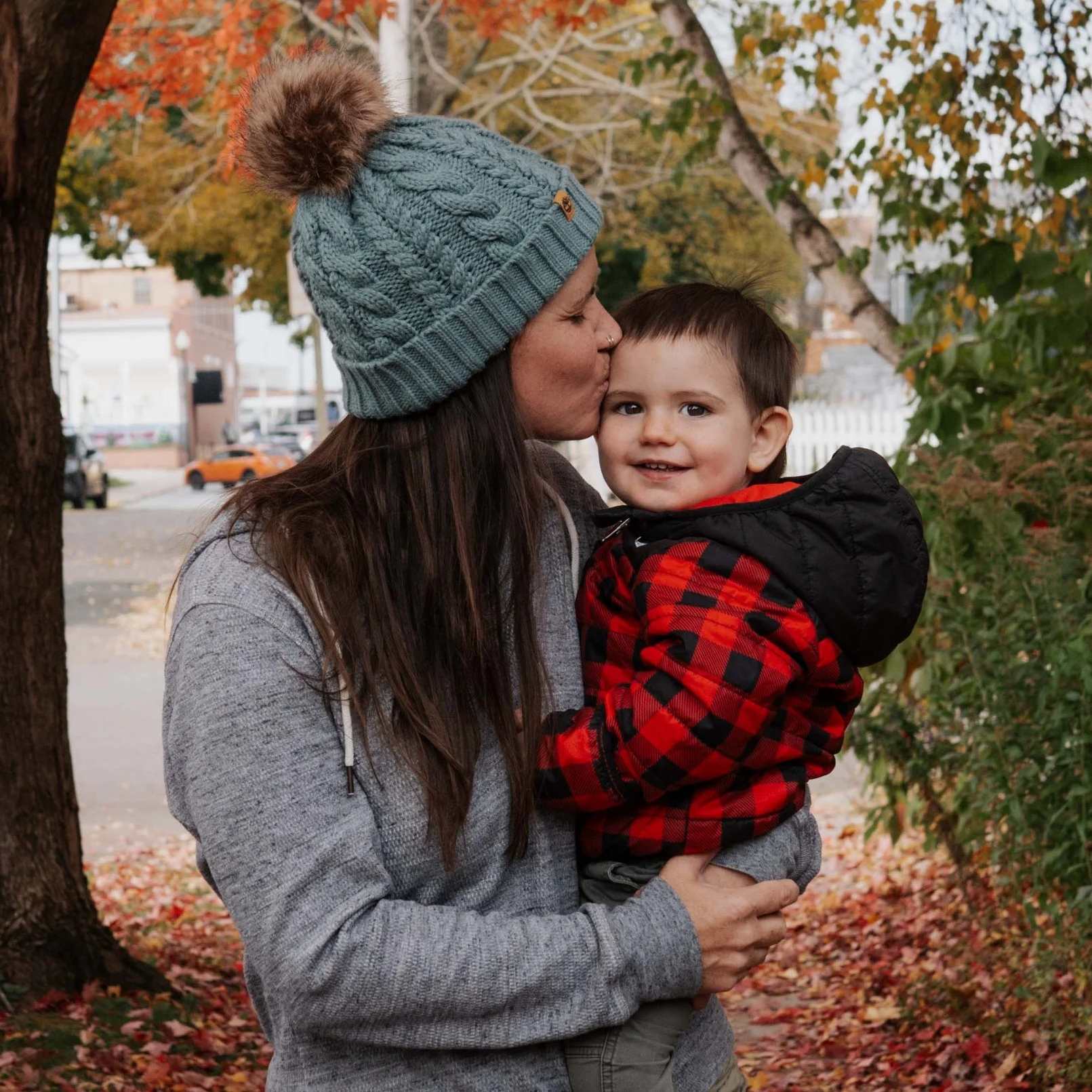 A woman in a gray hoodie and a gray knit cap with a pom-pom kisses a young boy on the cheek outdoors in autumn, with trees and fallen leaves in the background.