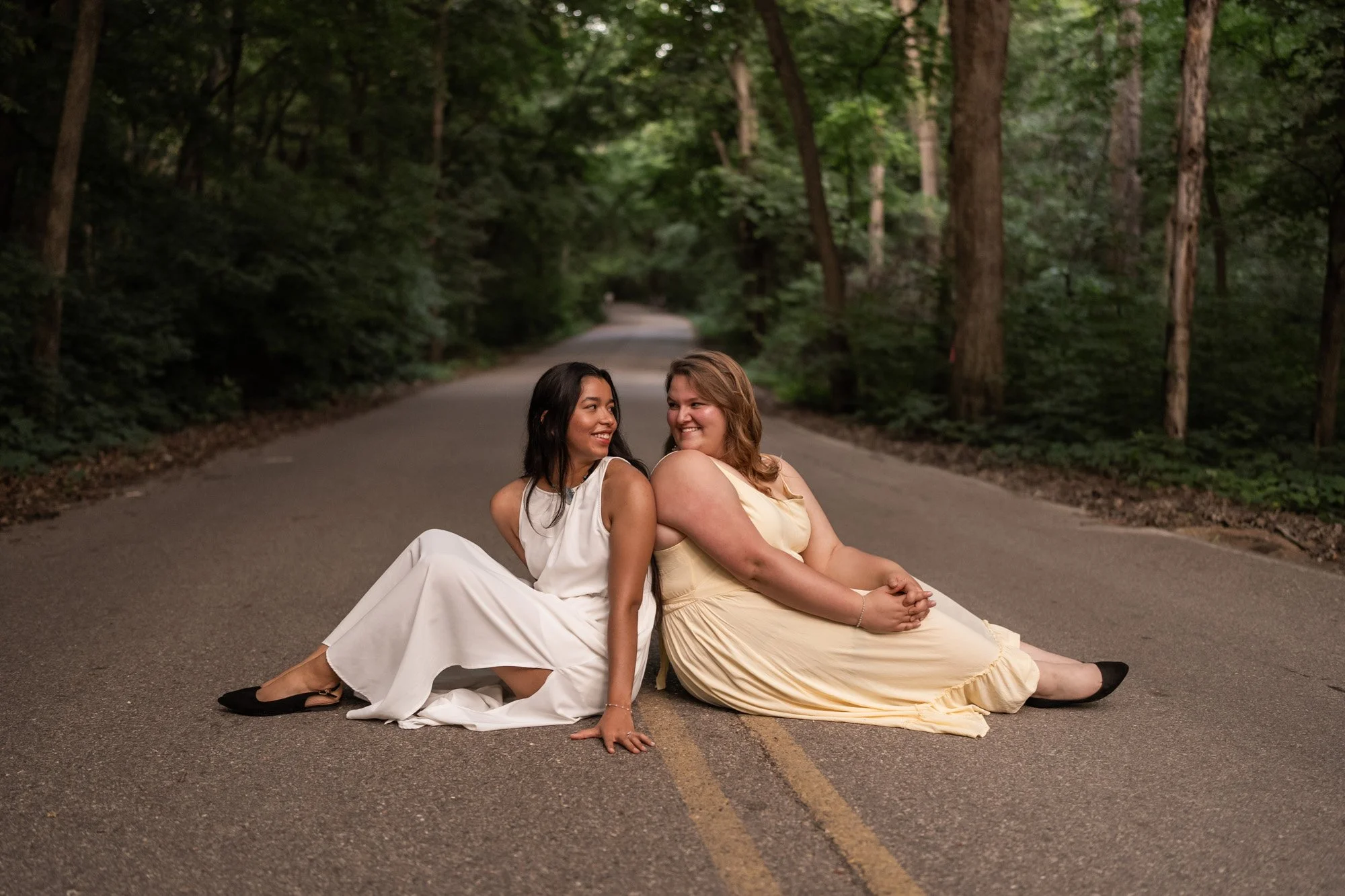 Two women in dresses sit on an empty forest road, smiling at each other, surrounded by tall trees and green foliage.