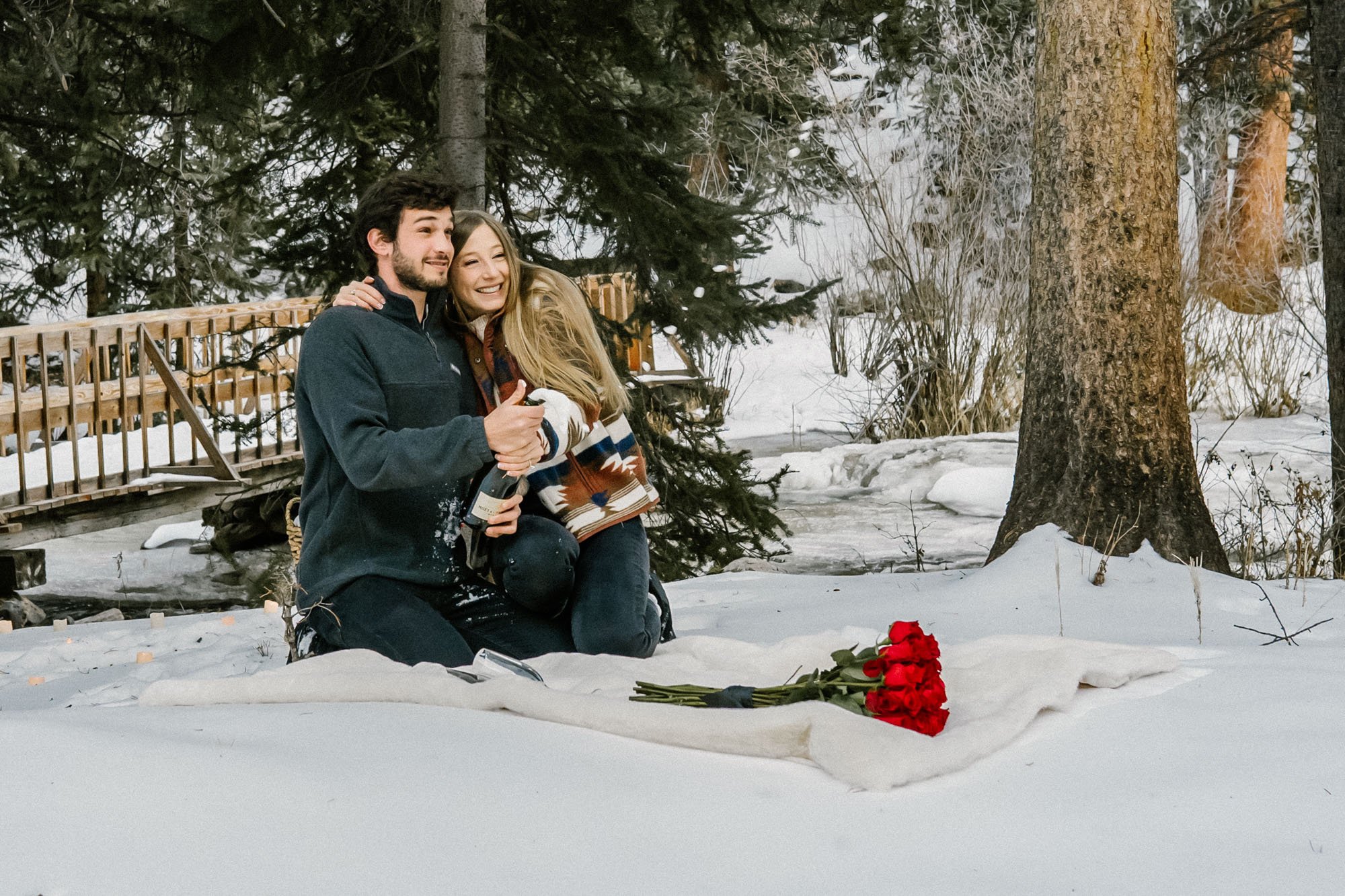 A couple celebrating a romantic moment post proposal, with a bouquet of red roses placed on a white cloth in front of them, outdoors surrounded by snow-covered trees and a wooden bridge in the background.