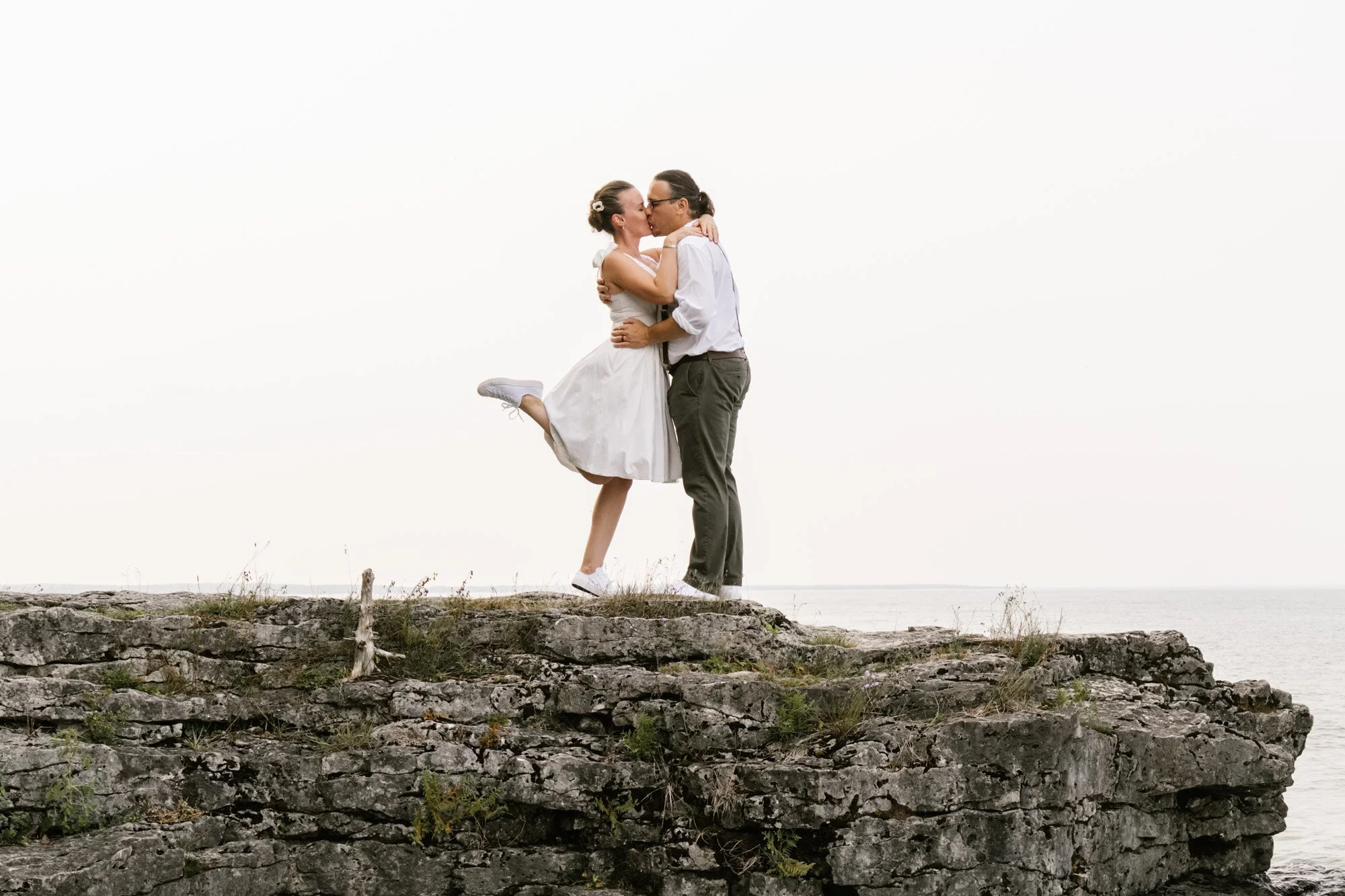 A couple kissing on a rocky cliff near the water, with the woman wearing a white dress and the man in a white shirt and dark pants, during daytime.