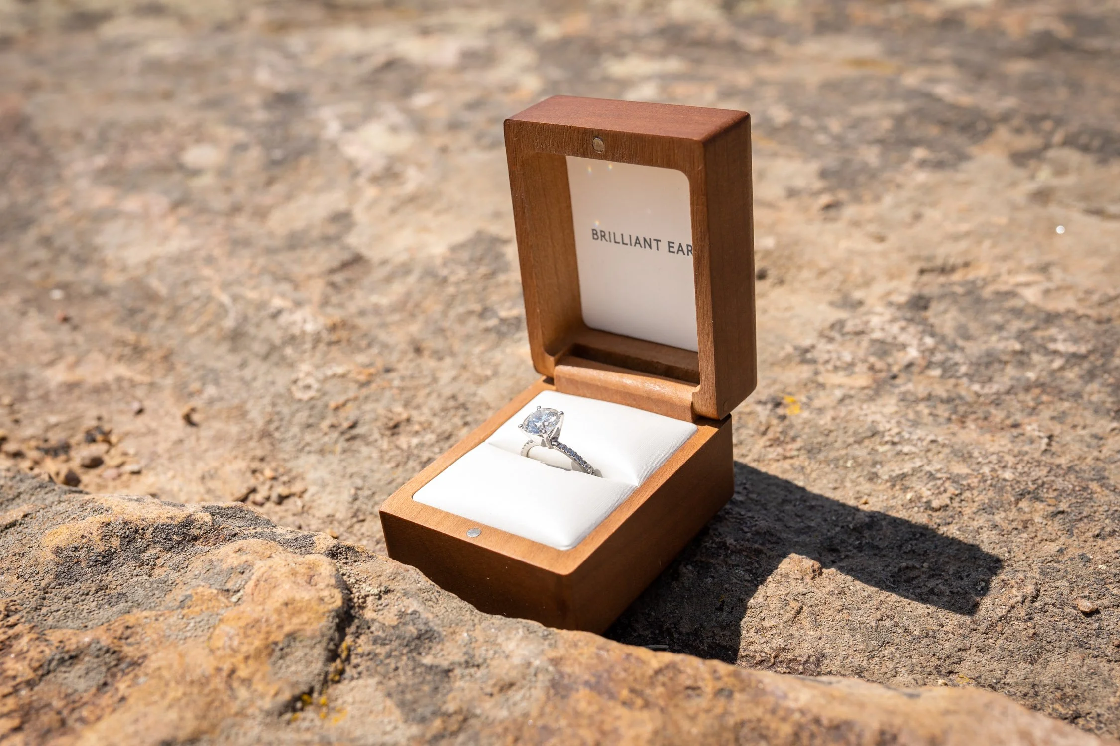 A diamond engagement ring in a wooden box on rocky ground with the inside of the lid reading 'Brilliant Ear'.