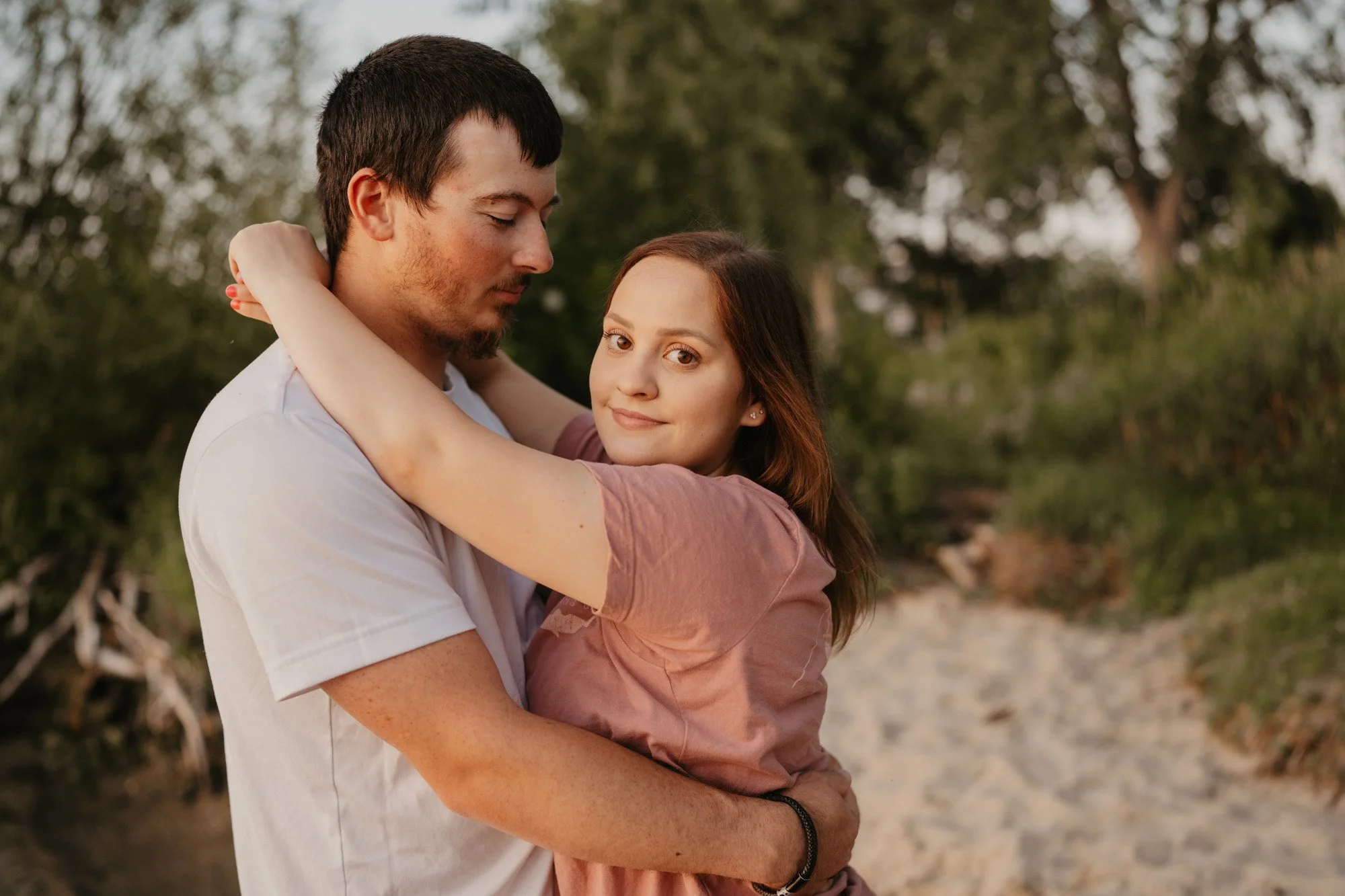 A young couple hugging outdoors on a trail with trees and greenery in the background.