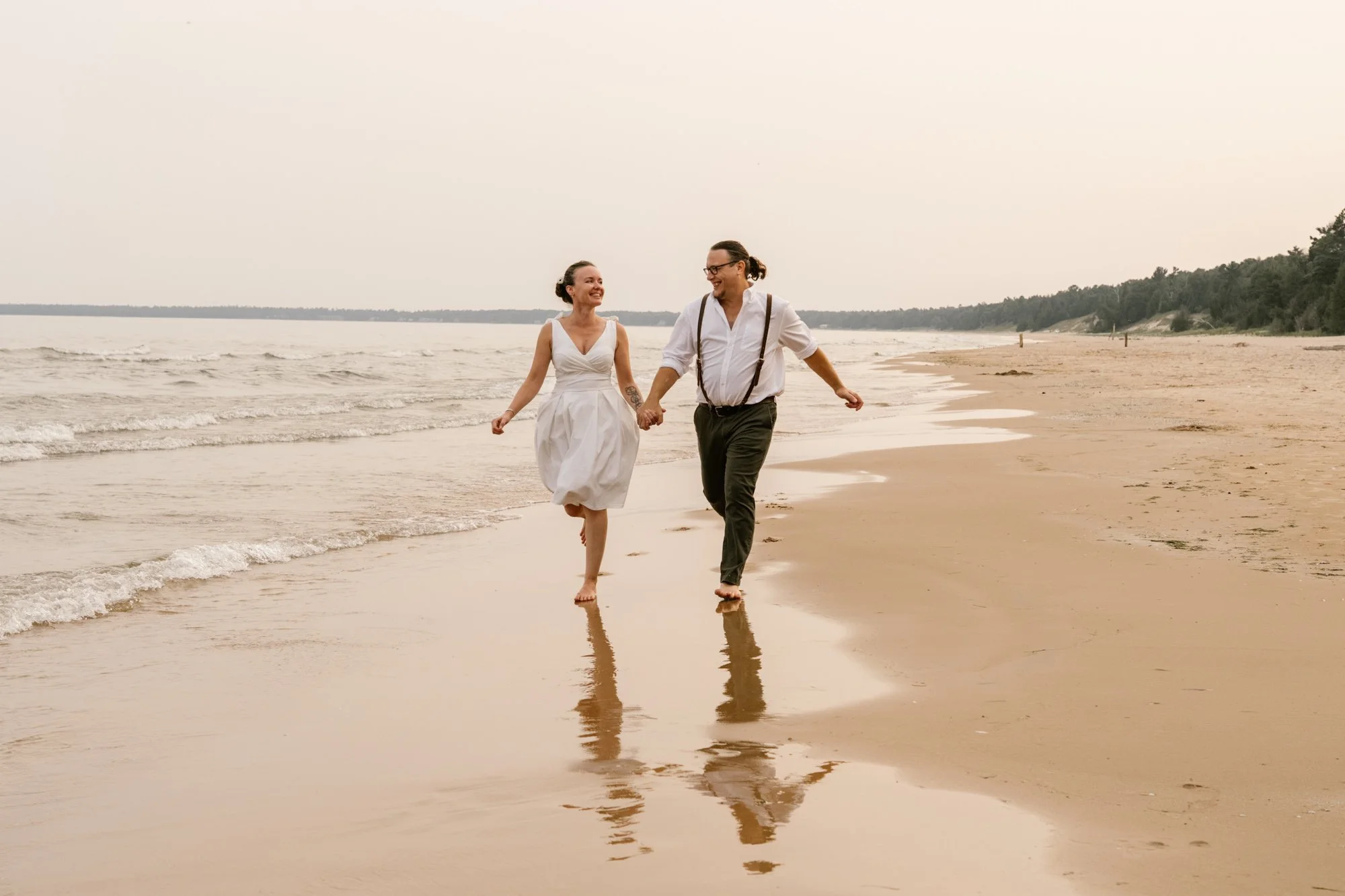 A couple holding hands and walking barefoot on the beach during sunset, dressed in vintage wedding attire, smiling and looking at each other.