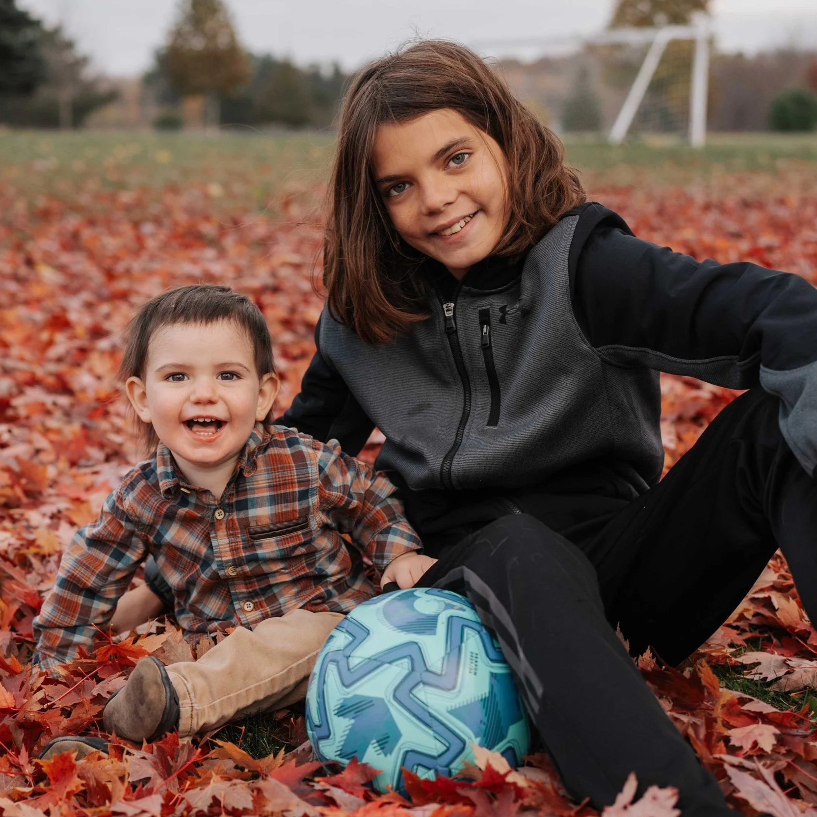 A boy and a toddler boy sitting on a bed of fallen autumn leaves with a soccer ball, smiling at the camera.