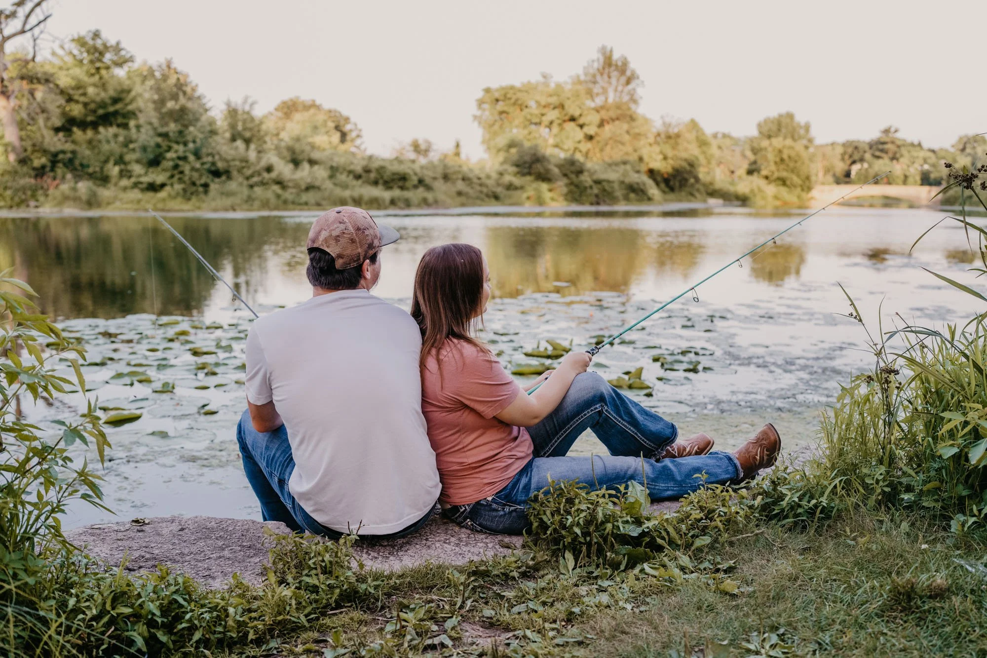 A man and woman sitting on a rock by a pond, fishing together during sunset.