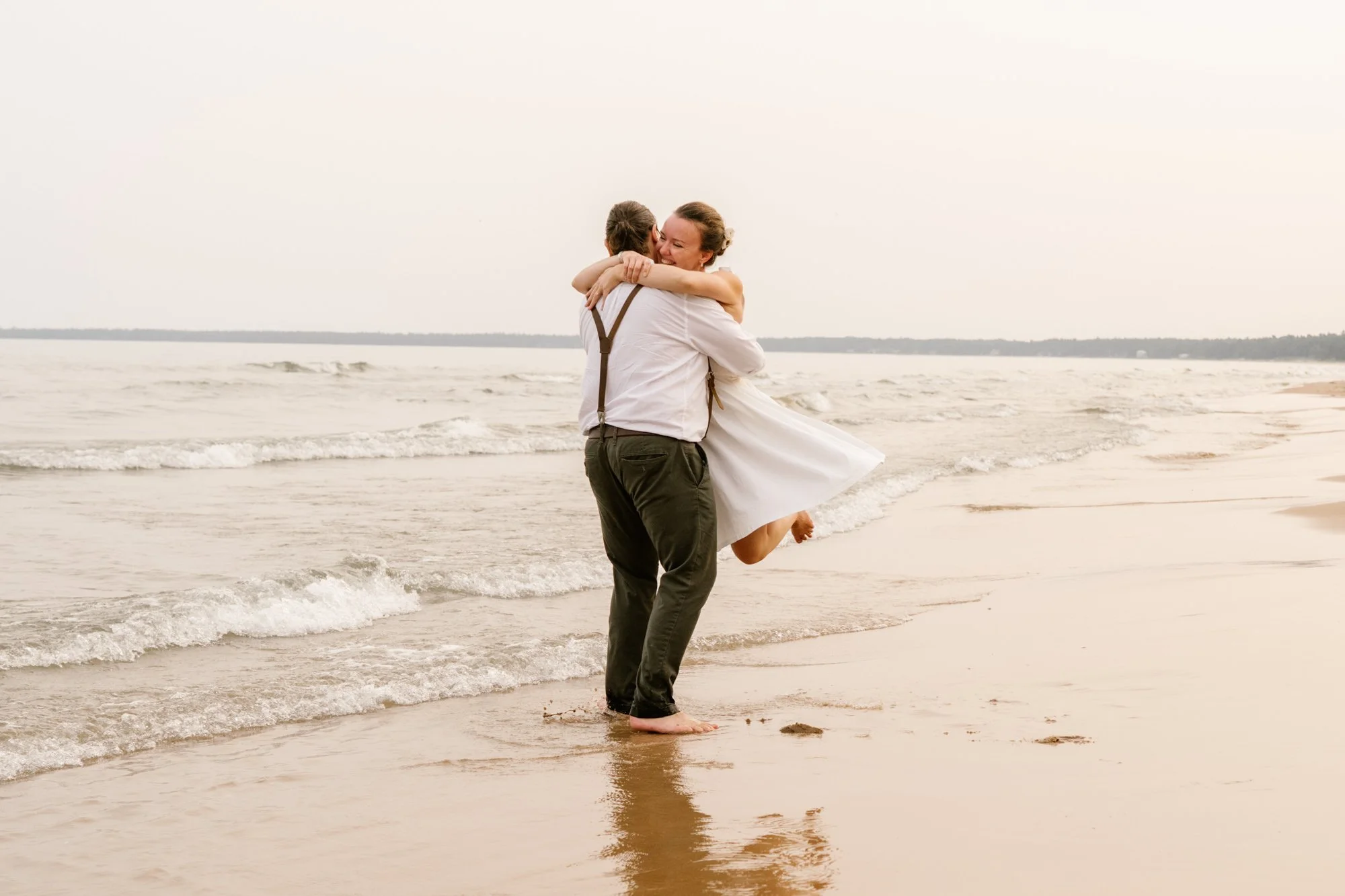 A couple on the beach, embracing and smiling, with the man lifting the woman in his arms; the woman is wearing a white dress, and the man has suspenders.