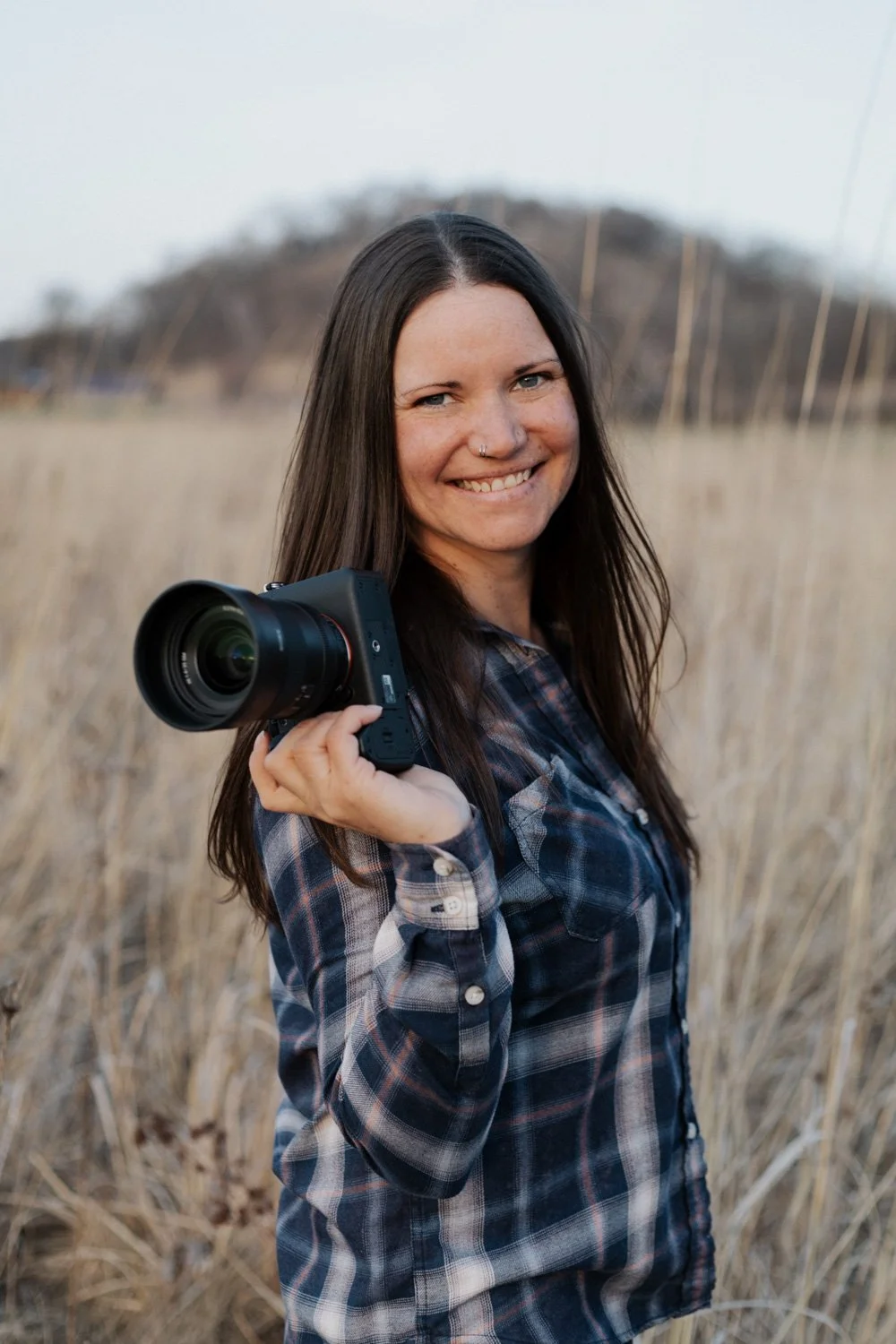 A young woman with long brown hair, smiling, holding a camera in a grassy outdoor field with hills in the background.