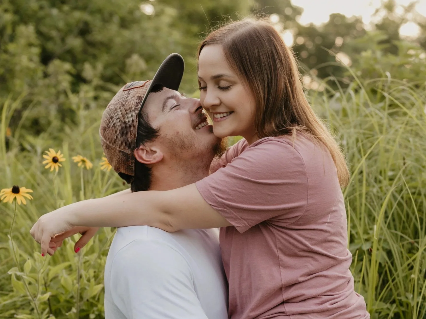 A couple outdoors in a grassy field with yellow flowers, smiling and hugging each other affectionately.