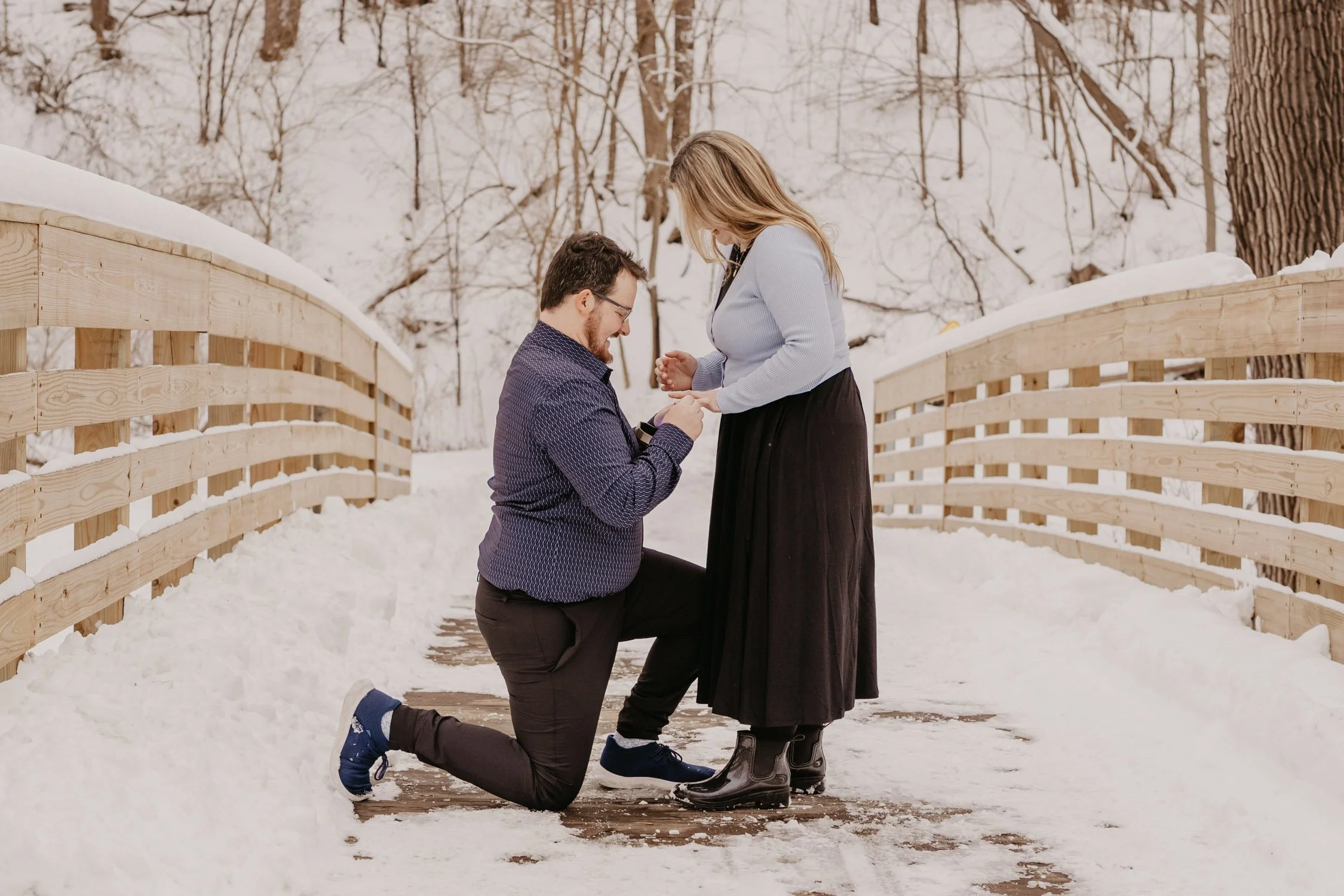 A man proposing to a woman on a snowy bridge in winter, with trees in the background.