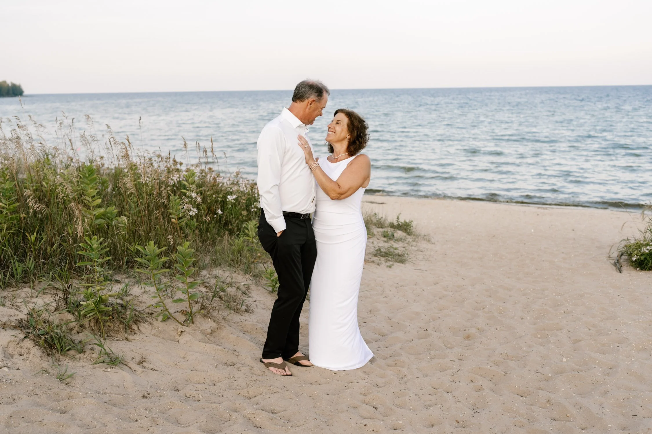A couple standing on a sandy beach, embracing and looking into each other's eyes, with the ocean in the background.