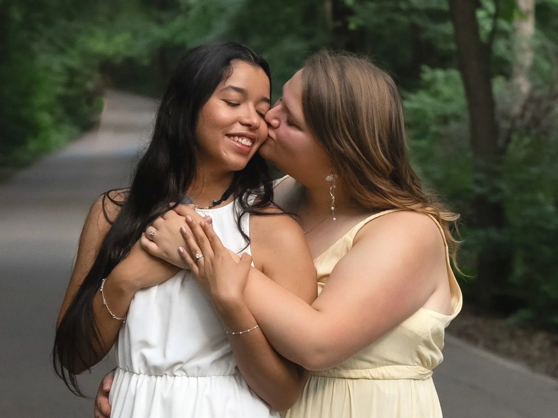 Two women embrace in a forested outdoor setting, one kissing the other's cheek with eyes closed, smiling.