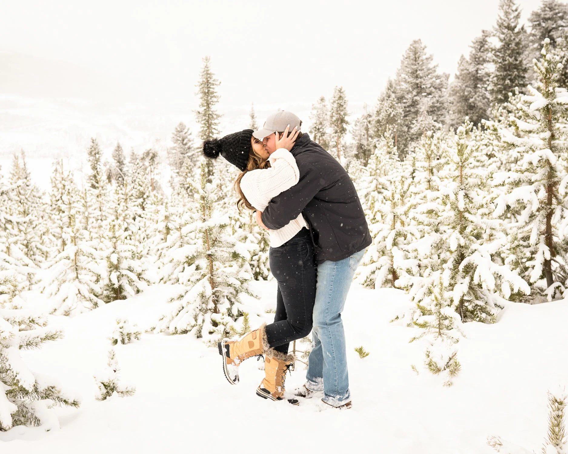 A couple in winter clothes kissing in a snowy forest with snow-covered trees.