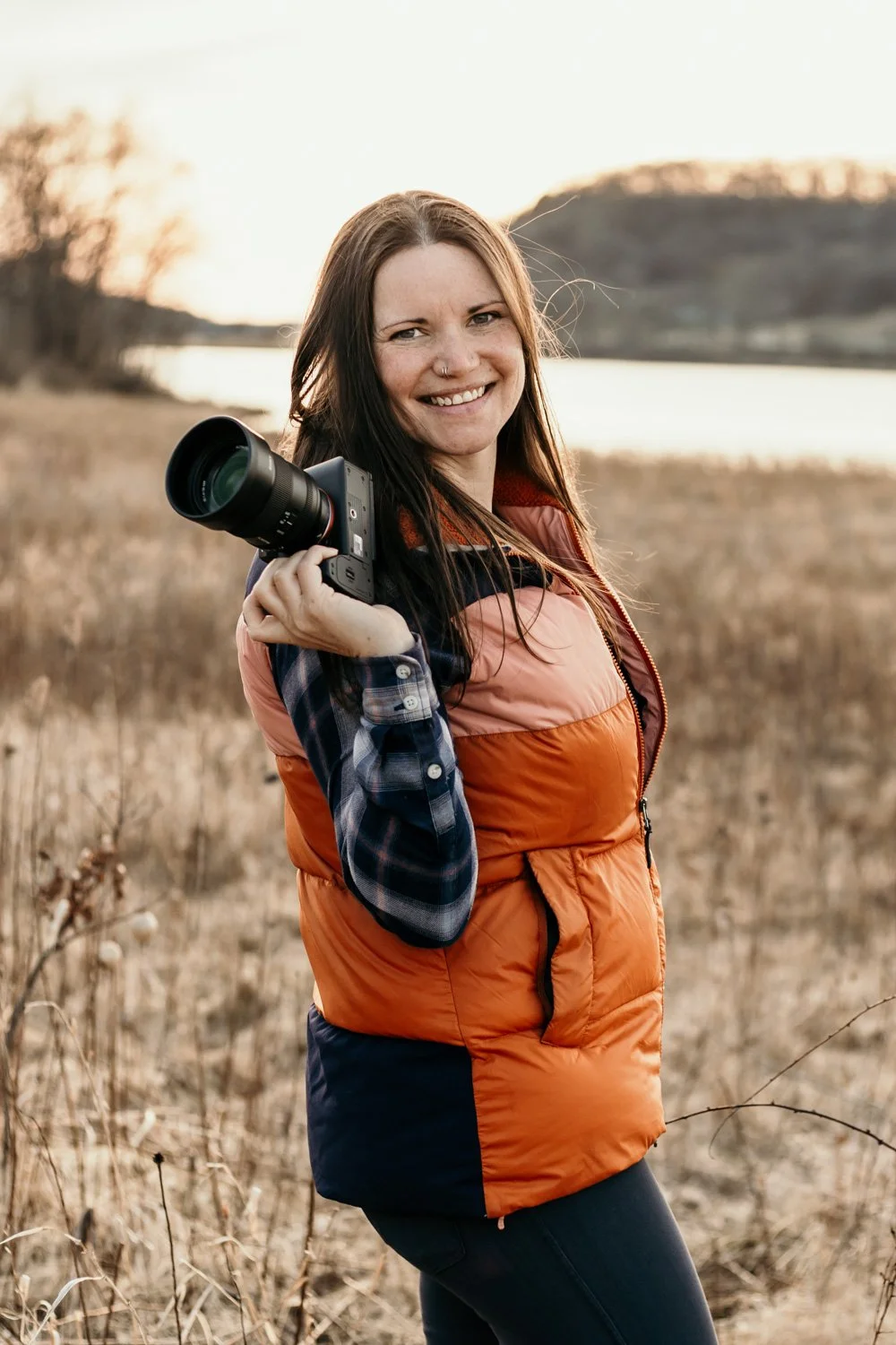 Woman smiling outdoors on a field with a camera over her shoulder, during sunset.