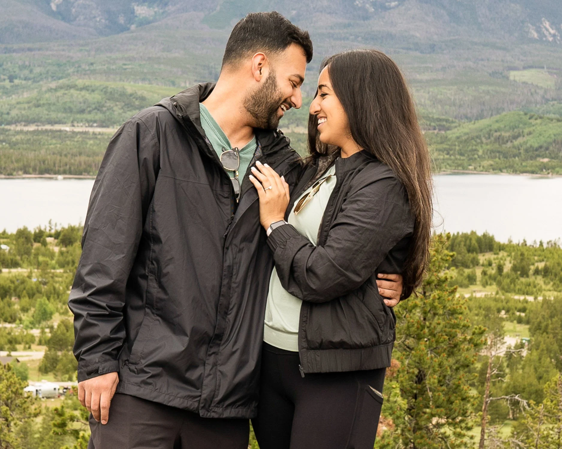 A happy couple standing close together outdoors with mountains, trees, and a lake in the background, sharing an intimate moment.
