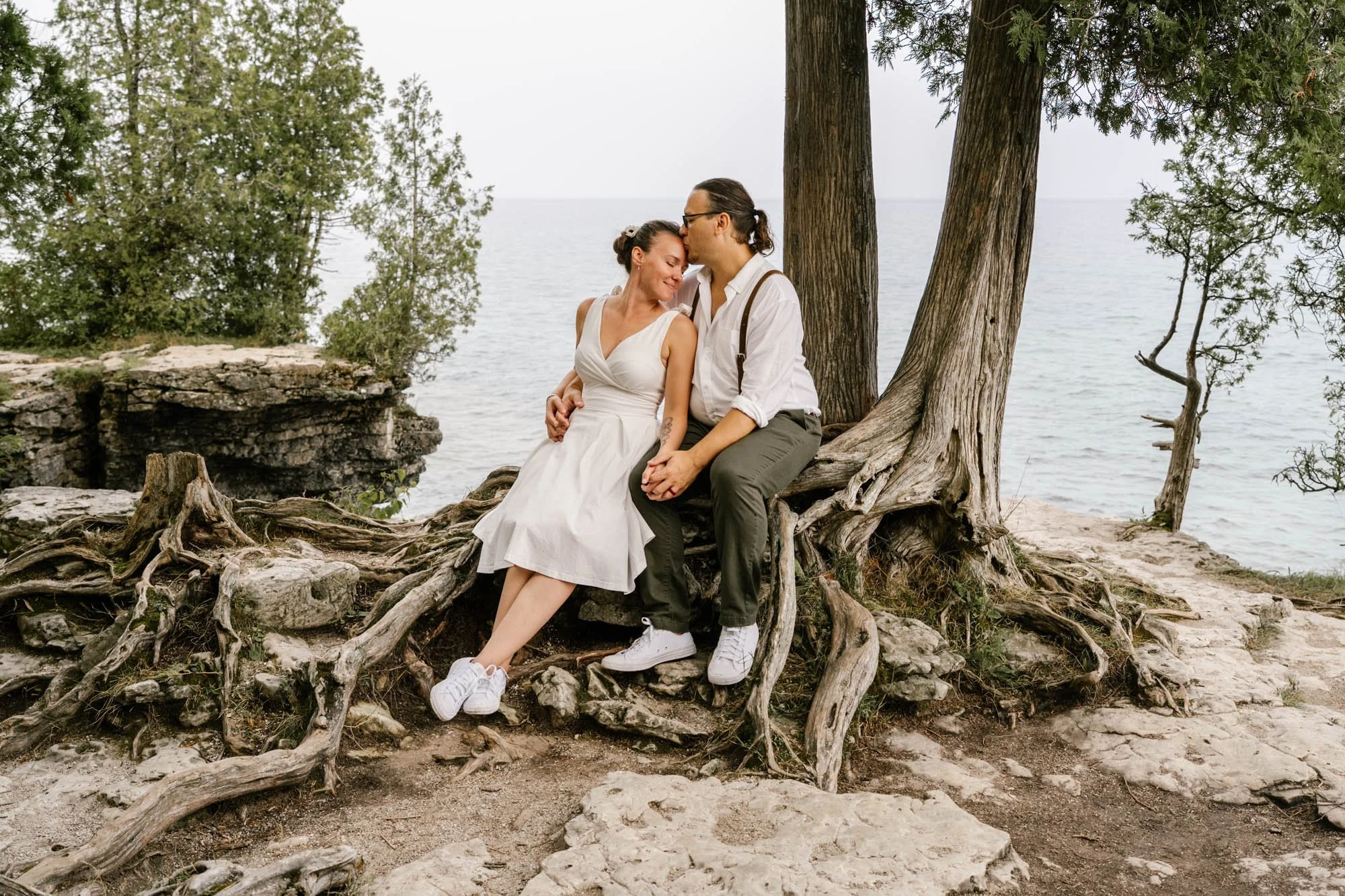 A couple sitting on a large tree root by the water's edge, sharing an intimate moment and kissing.
