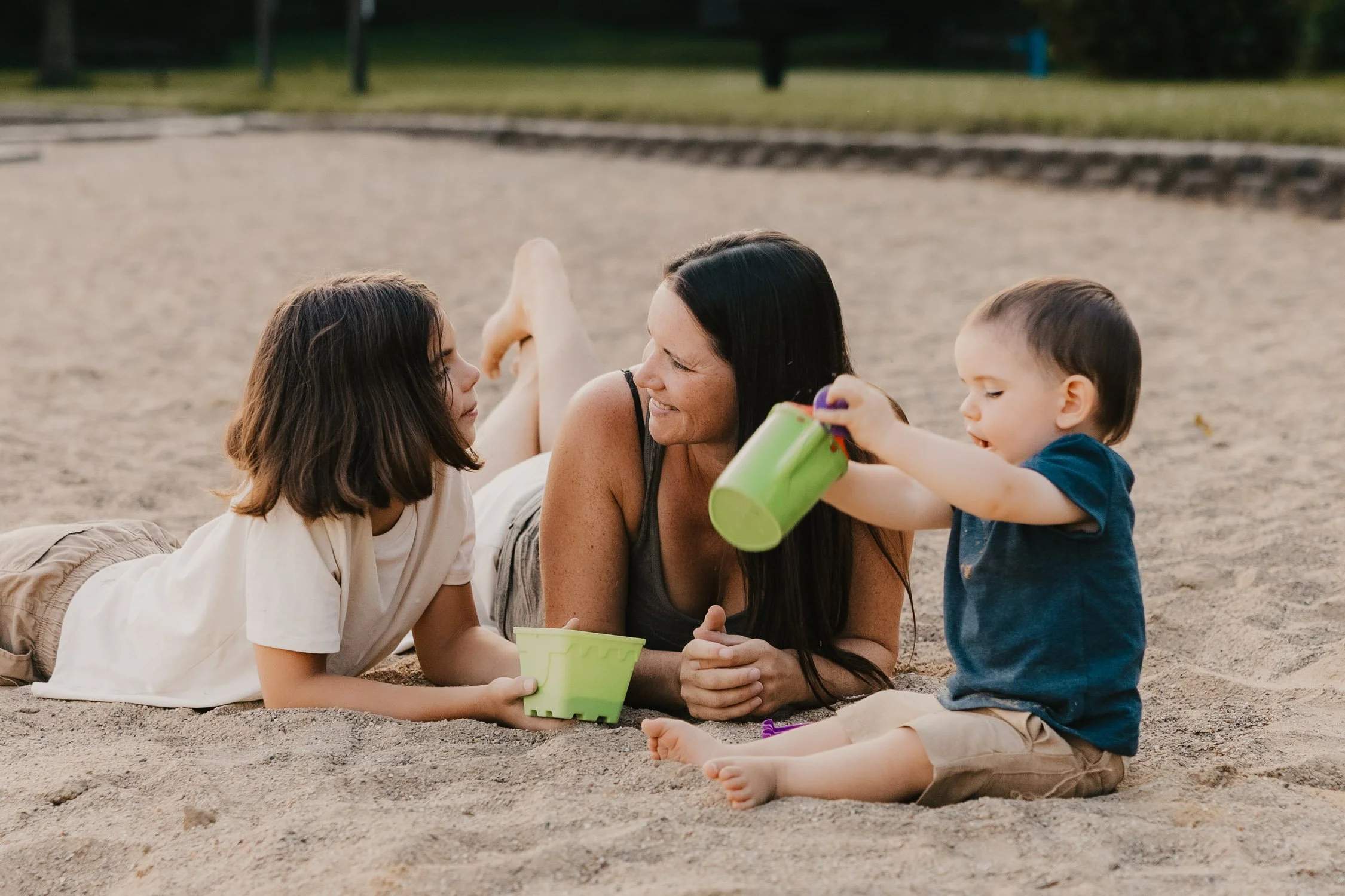A woman lies on her stomach on a sandy beach, smiling at a young girl with brown hair and a white shirt, while a young boy in a blue shirt and tan shorts plays with a green bucket nearby.