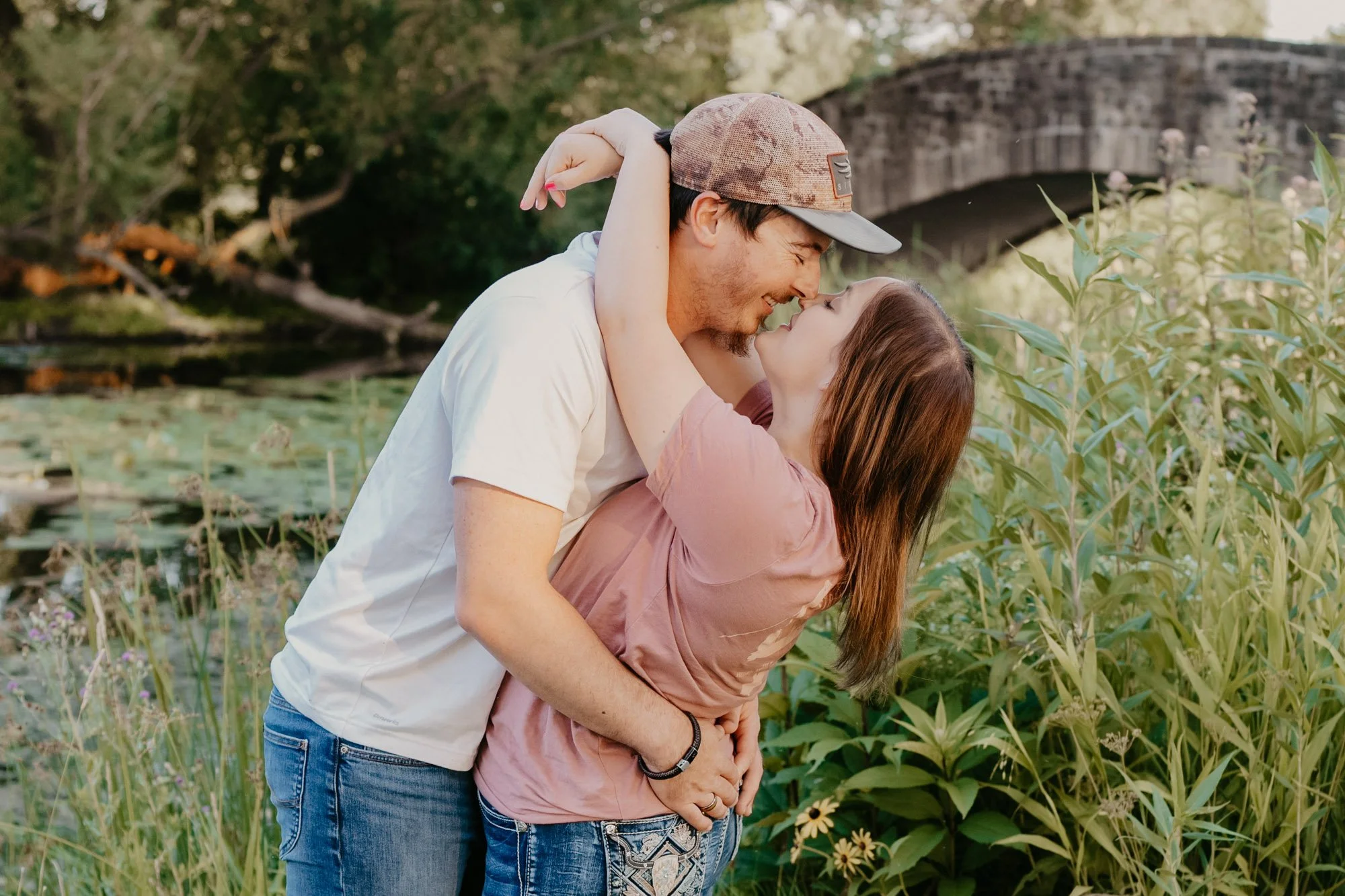 A couple embracing outdoors near a pond with lily pads, surrounded by tall green grass and plants, with a stone bridge in the background.